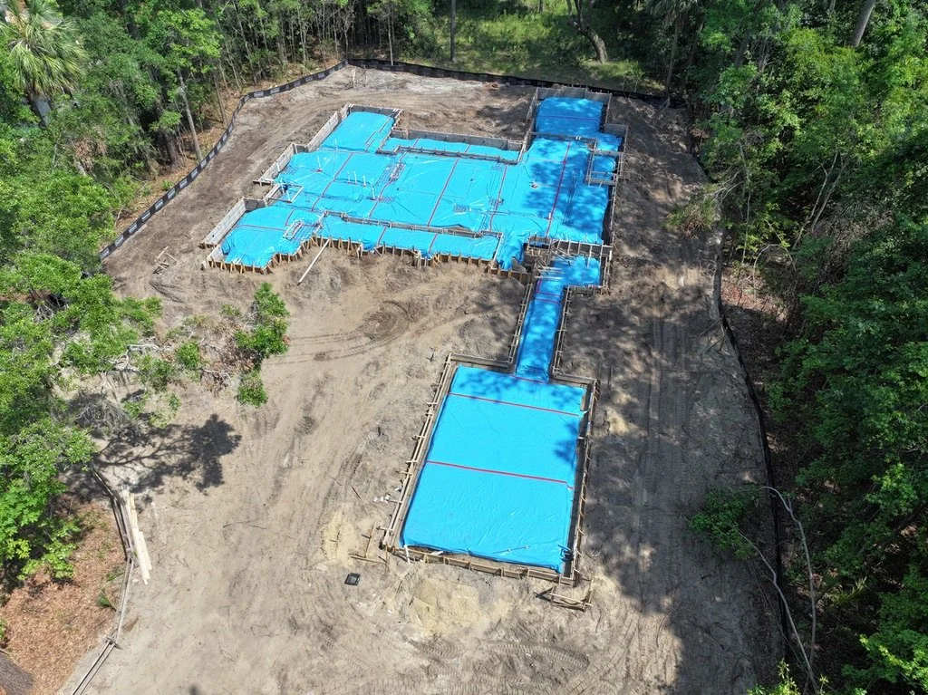 A construction site showing the foundation and footings of a building, with blue tarps covering parts of the structure, surrounded by trees.