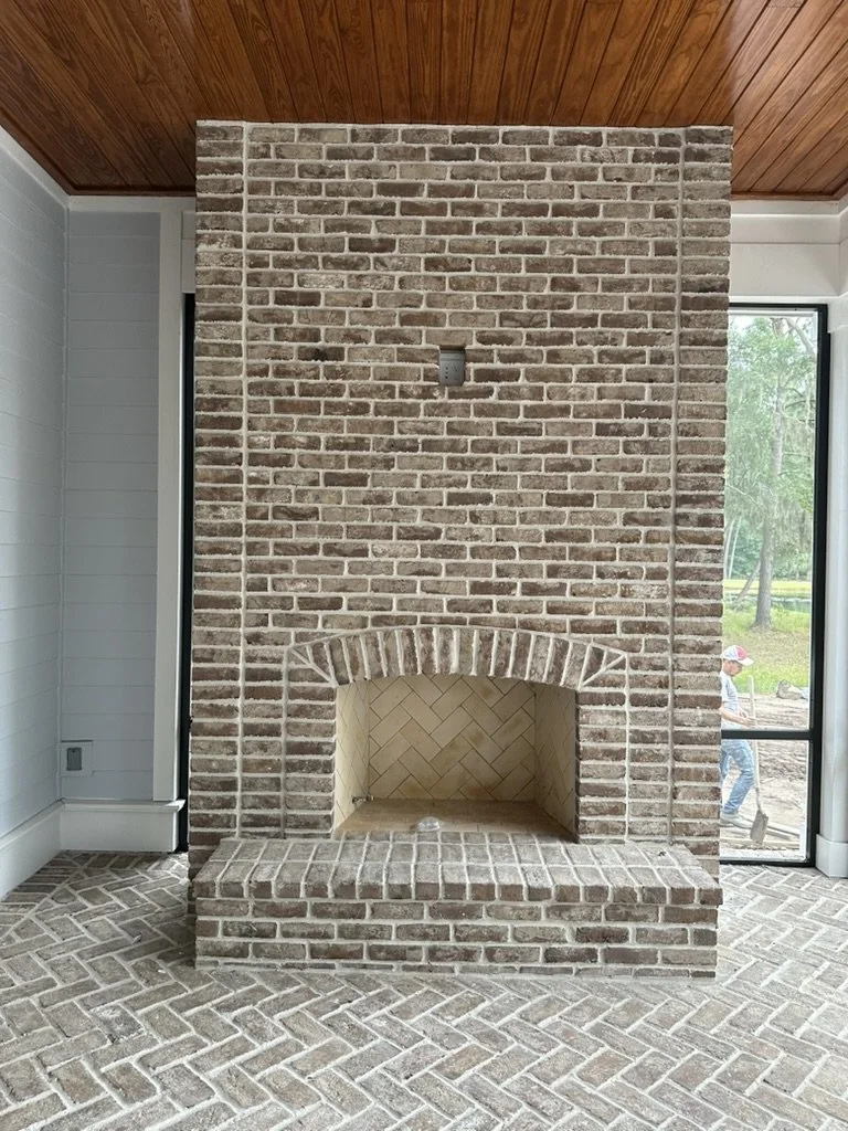 Brick fireplace with a herringbone-patterned interior, located in a room with a brick and herringbone brick floor and a wooden ceiling. A window and sliding door are on each side of the fireplace.
