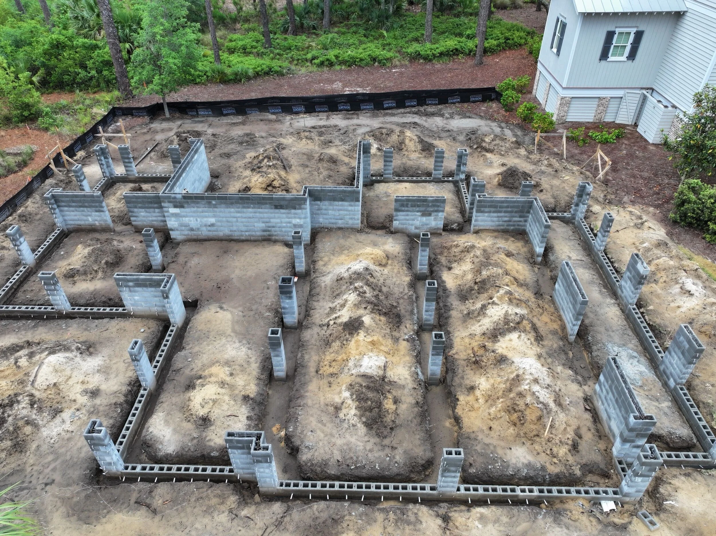 Foundation of a house under construction with cinder block walls and concrete blocks, surrounded by soil and dirt, with green trees and a house in the background.