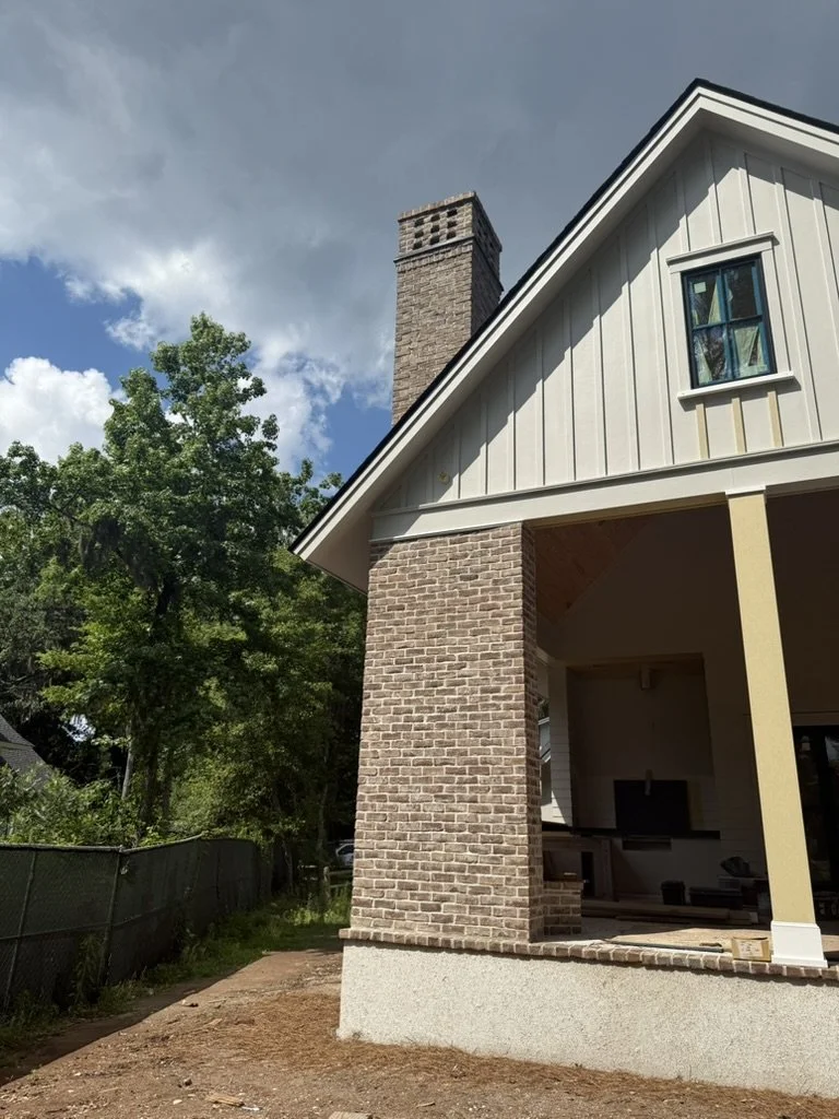 Side view of a house with a brick chimney, white siding, and a covered porch area, with a green tree and partly cloudy sky in the background.