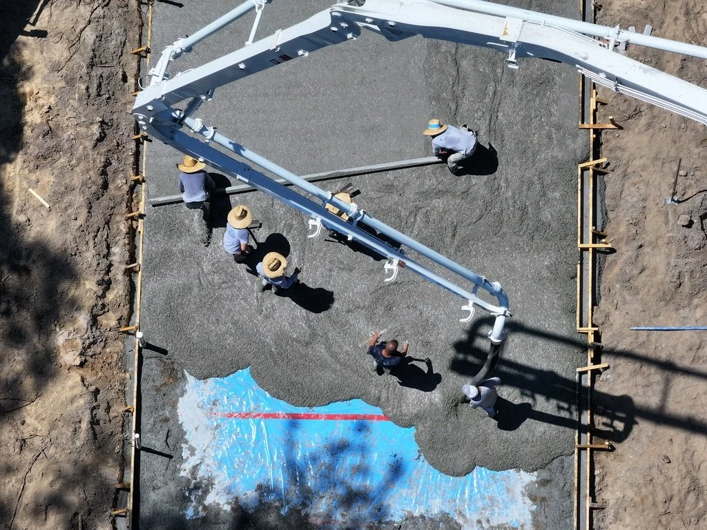 Construction workers pouring concrete into a foundation, with a partially finished area showing a painted water body outline, surrounded by dirt and construction equipment.