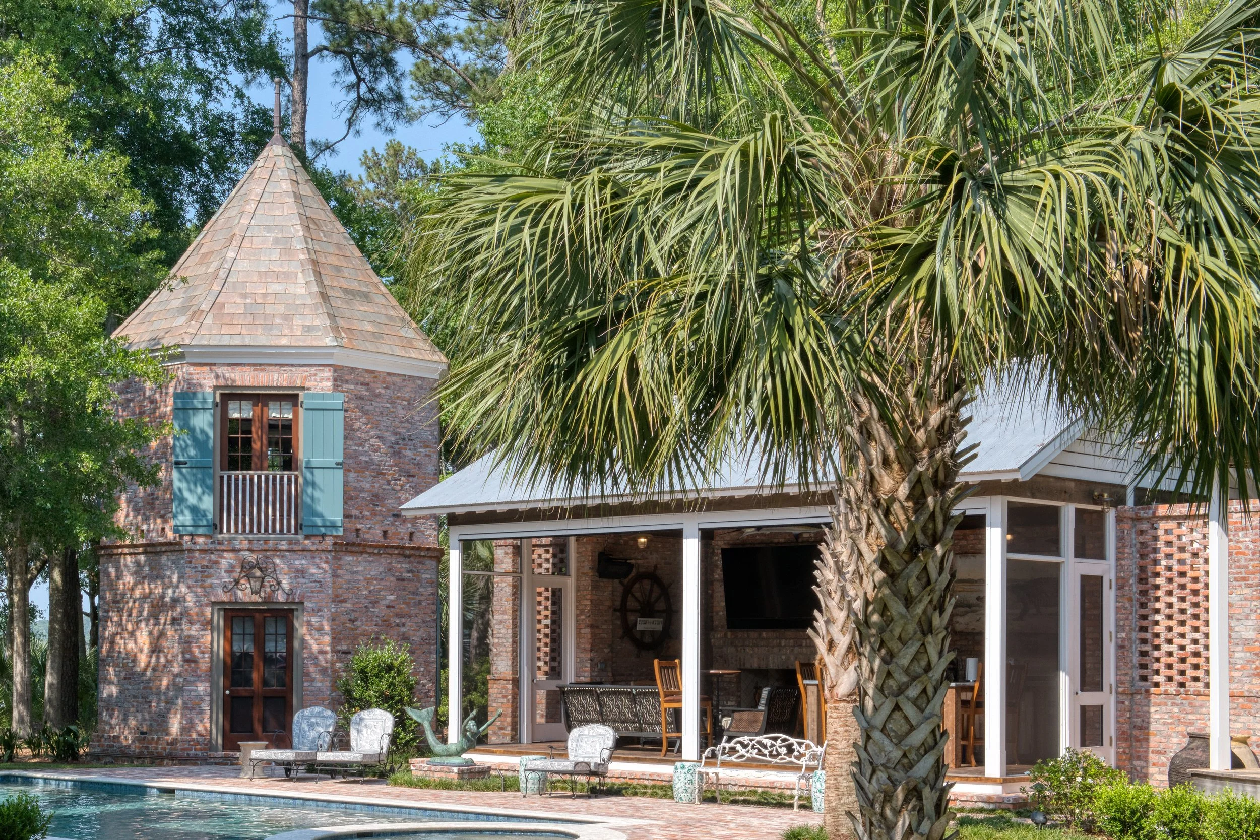 A backyard with a swimming pool, a brick tower with blue shutters, and a screened porch with outdoor furniture, trees, and a large palm tree.