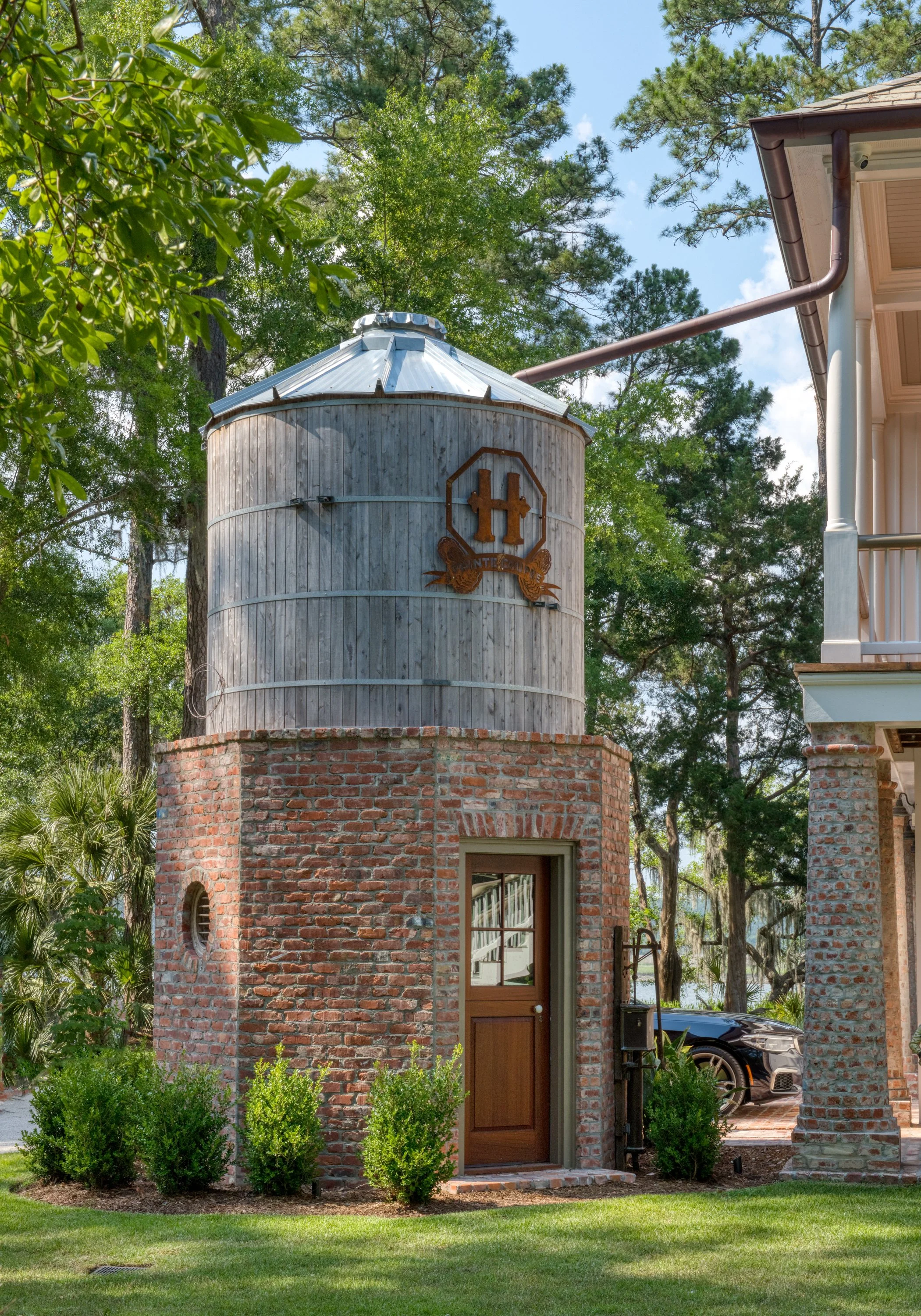 A small brick and wood building with a galvanized metal silo on top, featuring a door and small window, surrounded by greenery and trees, with a car parked nearby.