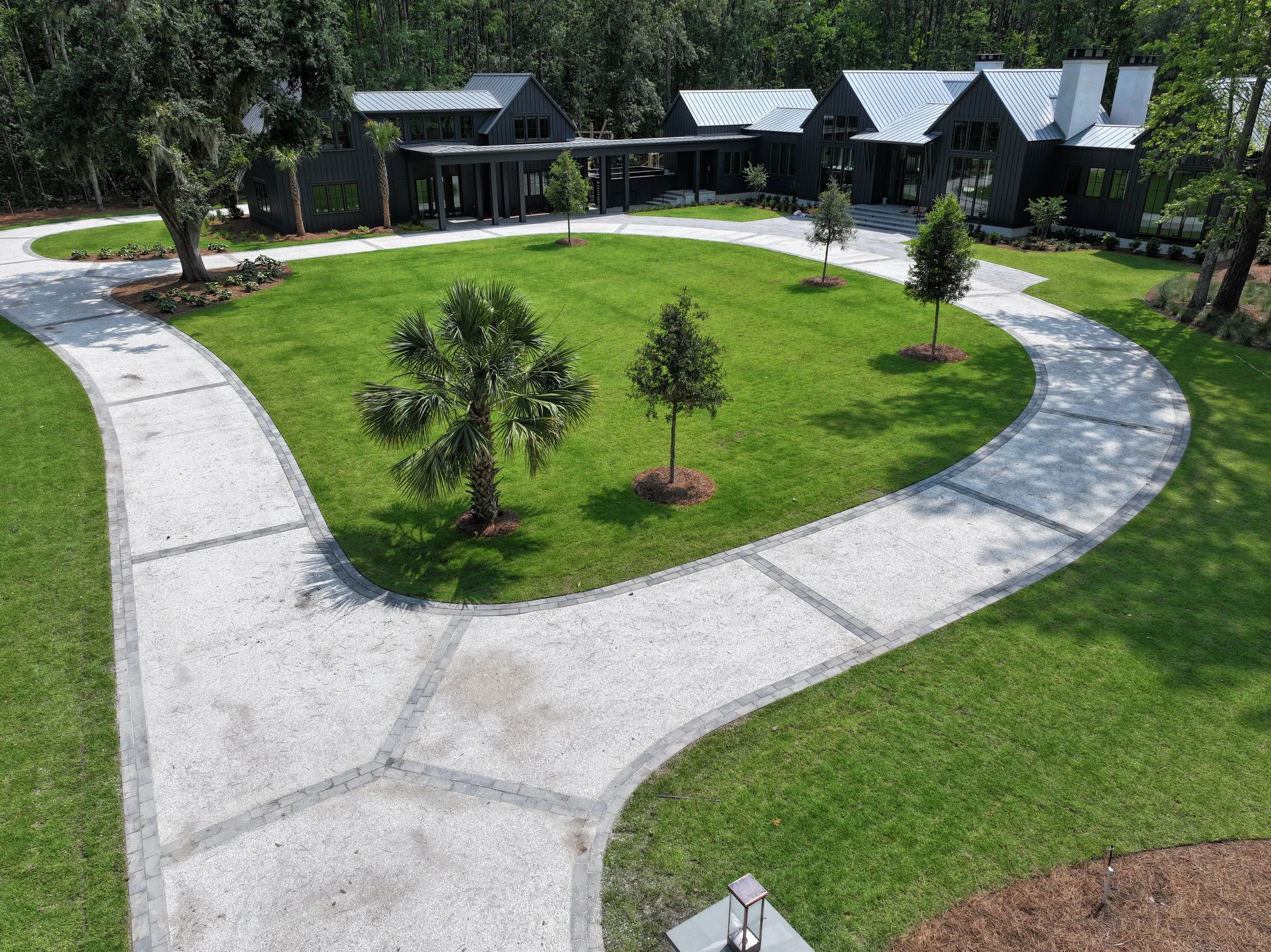 A modern house with dark exterior walls and large windows, surrounded by a landscaped yard with grass, small trees, and a curving paved walkway.