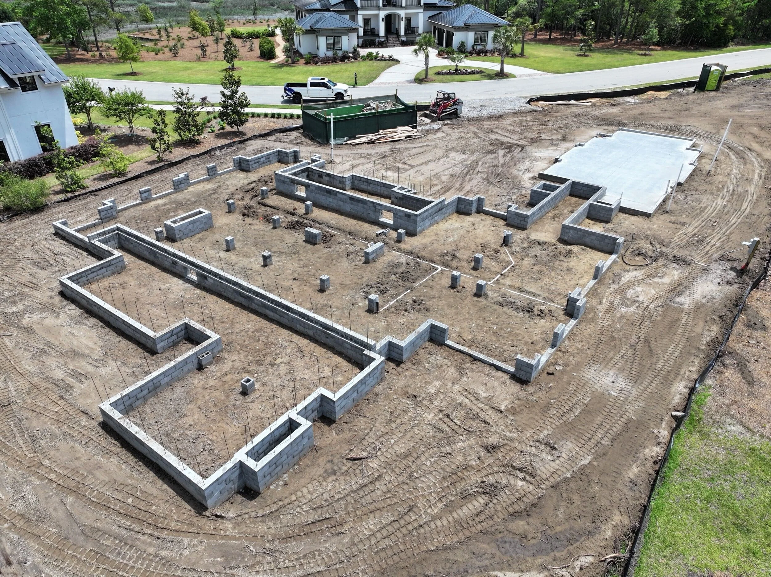 Under construction house foundation with concrete blocks, metal rods, and part of concrete slab in a suburban neighborhood with completed houses, trees, and a vehicle in the background.