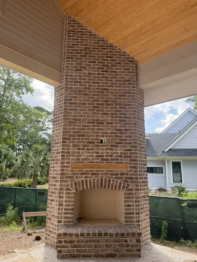 Brick outdoor fireplace with arched opening, situated outside a house with white siding and neighboring trees.