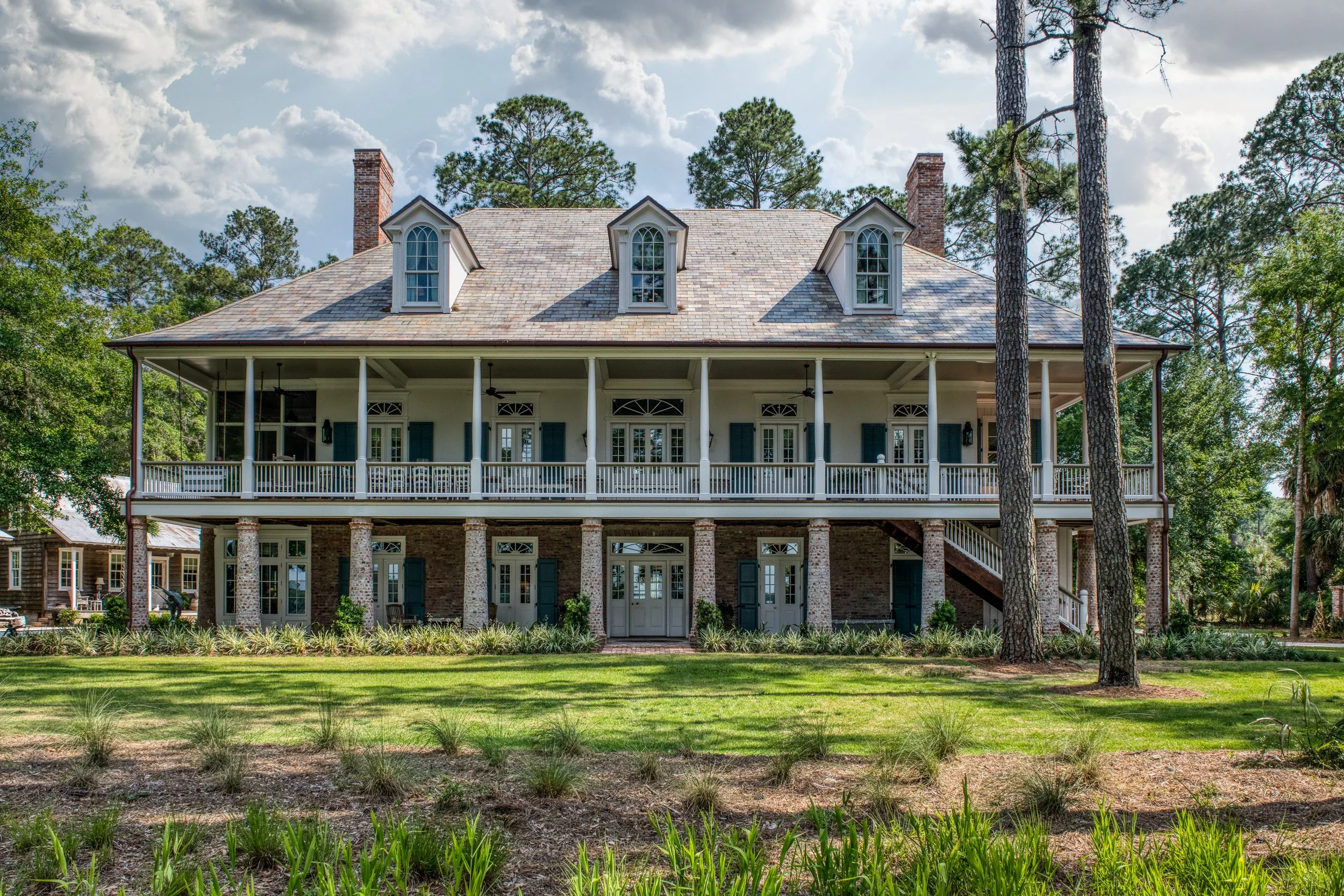 Large two-story house with brick and white siding exterior, tall trees, a spacious porch with white railings, multiple doors and windows with shutters, three dormer windows on the roof, and a well-maintained yard with grass and plants.