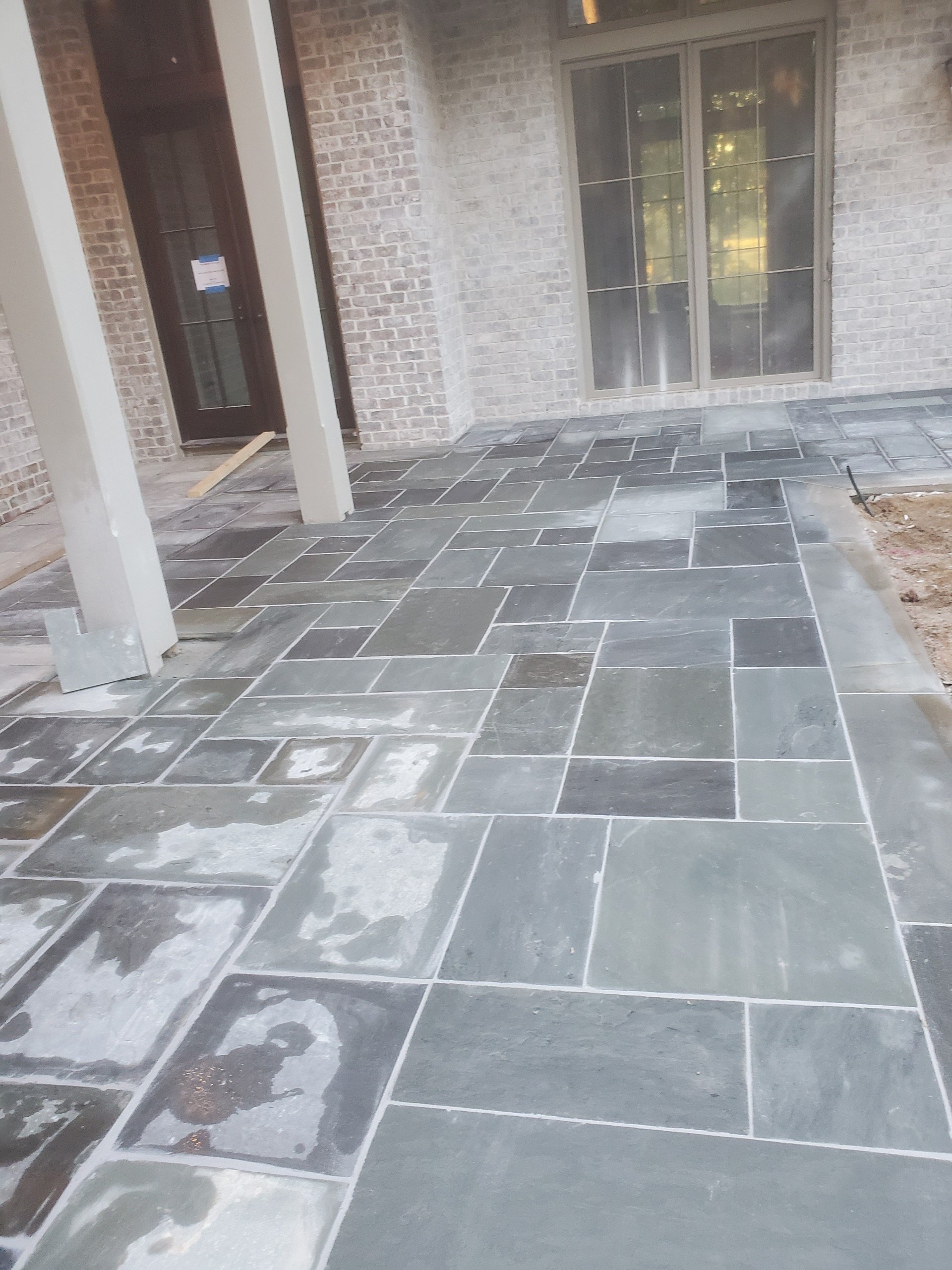Front porch with tiled stone flooring and brick exterior wall of a house, featuring a sliding glass door and a front door.
