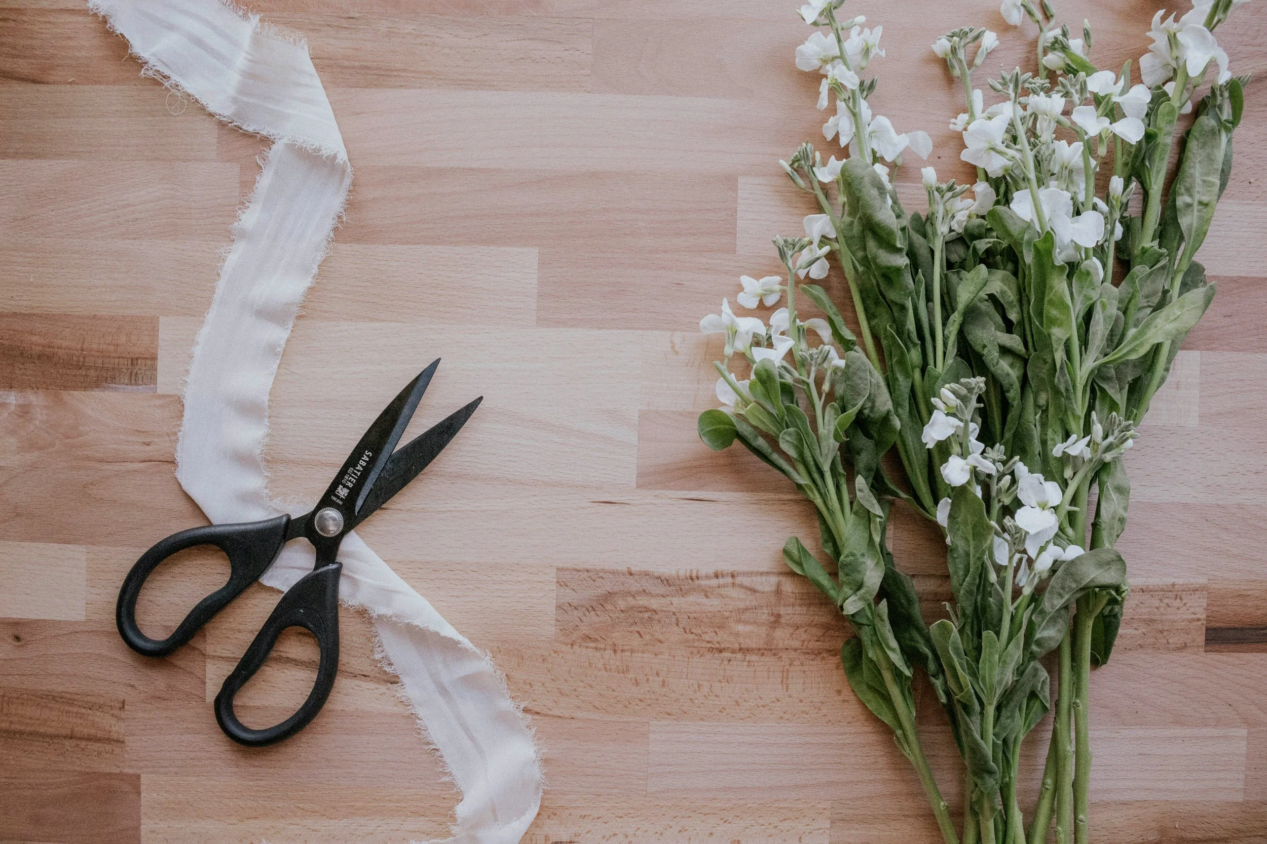 A bundle of white flowers with green leaves on a wooden surface, a pair of black scissors, and a piece of white ribbon.