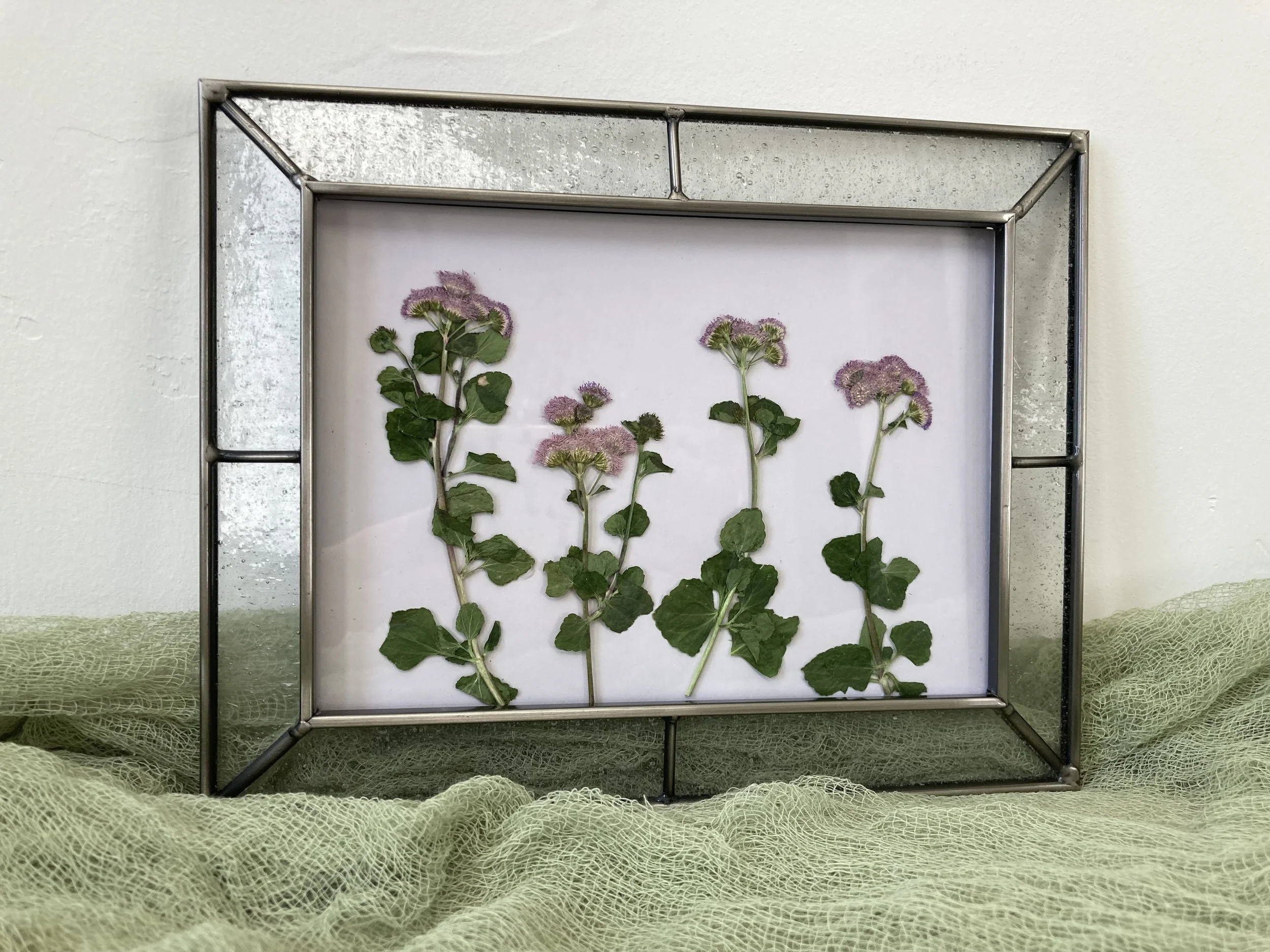 Framed botanical artwork featuring pinkish-purple flowers with green leaves on a white background, placed on a textured green surface.