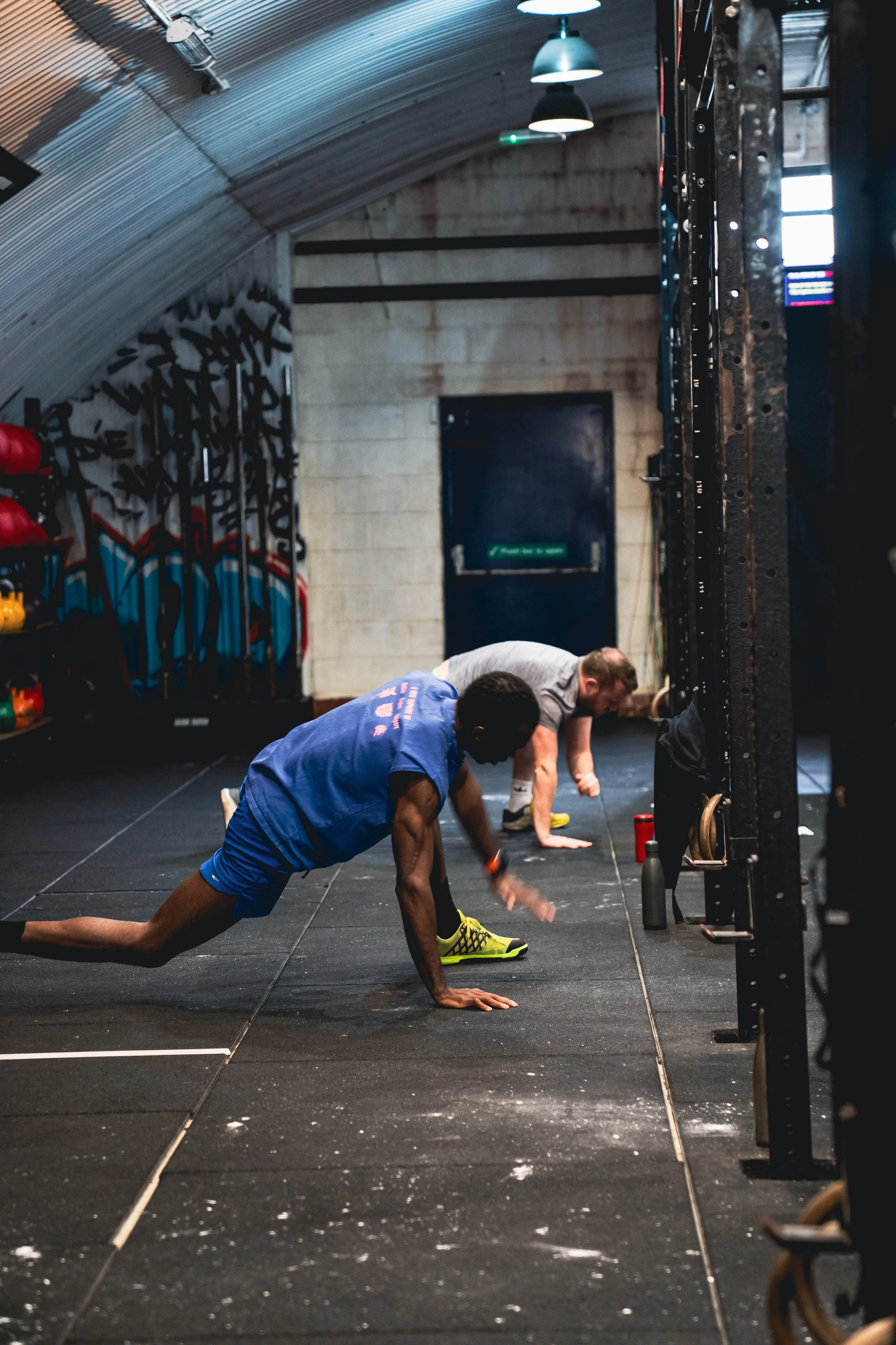 Two men doing push-ups on a black gym floor inside a gym with equipment visible on the sides.