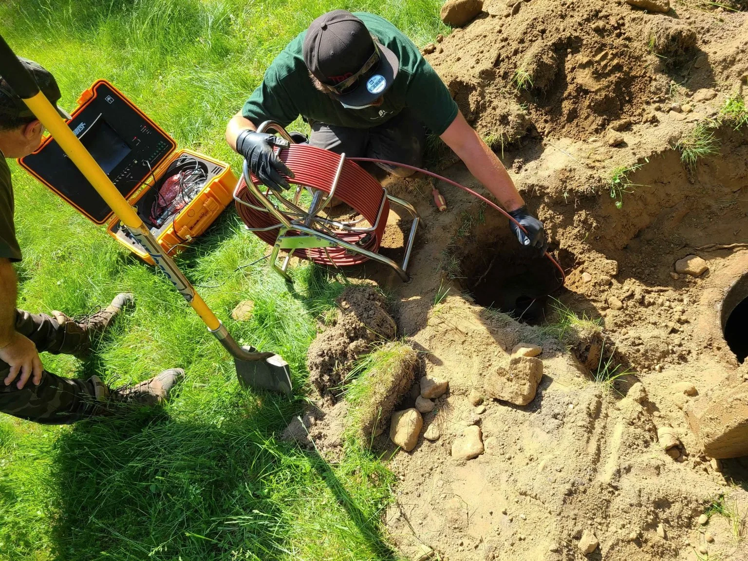 Deux personnes travaillent dans un trou dans le sol, utilisant un équipement de détection géologique, avec une boîte à outils à proximité. Ils se trouvent dans un espace en plein air avec de l'herbe verte autour.