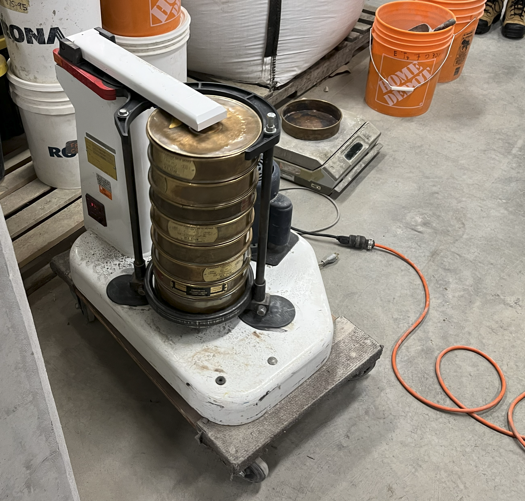 Une machine de pesée de conserve, probablement utilisée pour la mise en boîte d'aliments, dans un environnement industriel ou de stockage avec des seaux orange et blancs de marque Home Depot autour.