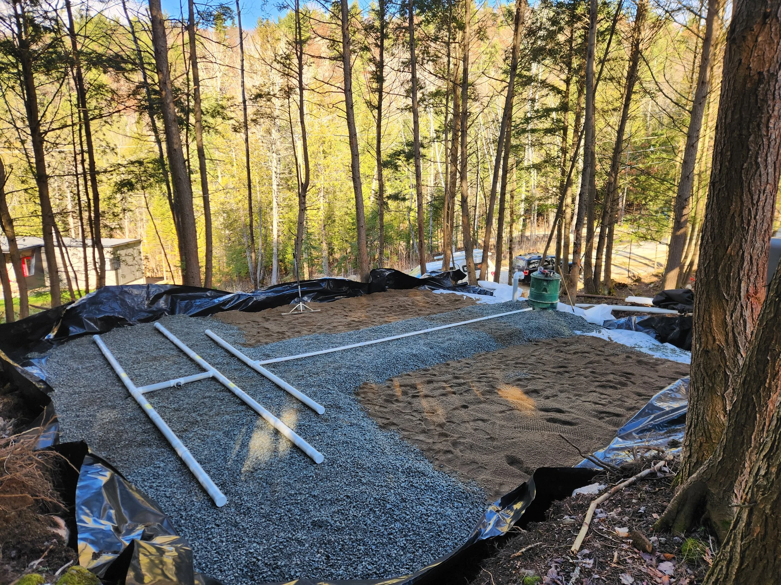 Pose de fondations pour une construction dans une zone boisée, avec des tuyaux en PVC alignés sur une surface recouverte de gravier, entourée d'arbres.