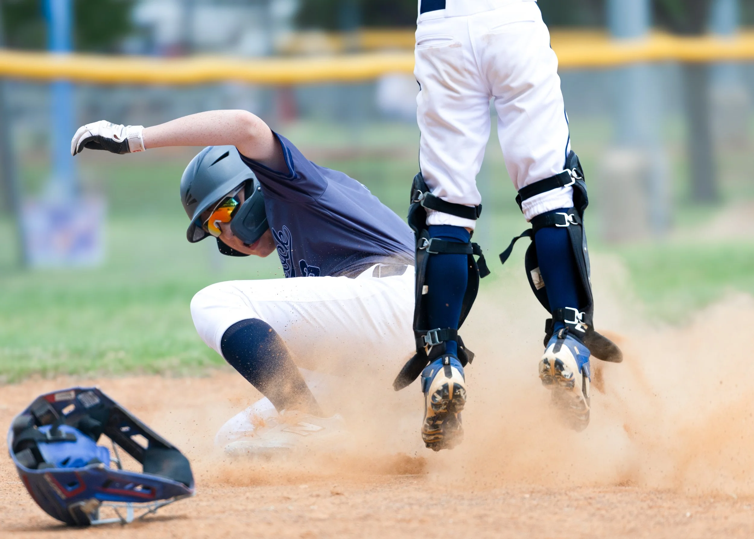 A baseball player in a navy jersey and white pants kneels on the ground after sliding into a base, with a teammate standing nearby on the dirt field. The player is wearing a helmet and sunglasses, and there is a baseball glove on the ground.