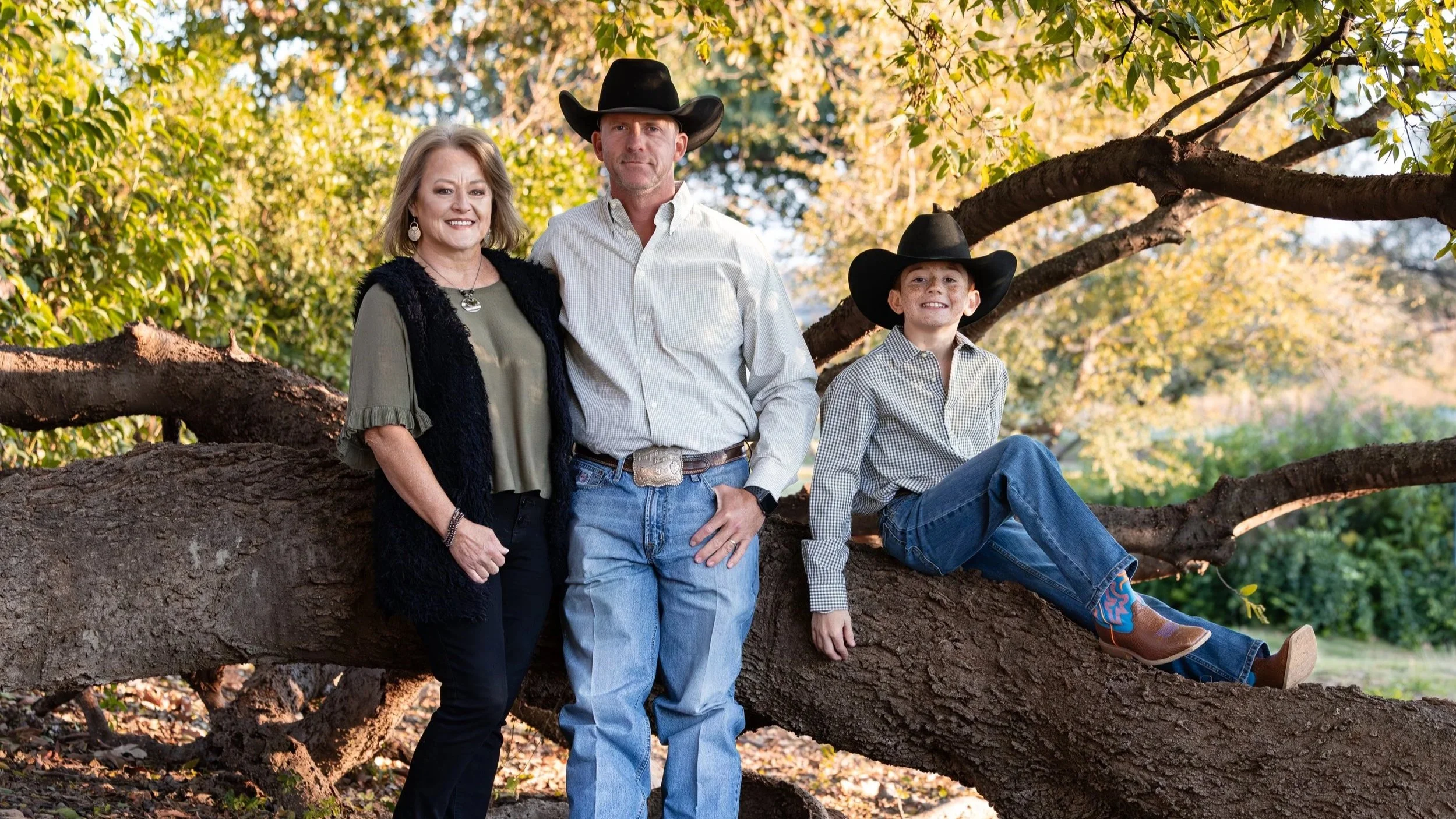 A family of three dressed in western attire, posing outdoors beneath a large tree with sprawling branches. The woman is smiling, wearing a green blouse, black vest, and jewelry. The man stands next to her with one hand in his pocket, wearing a white shirt, jeans, and a cowboy hat. The boy is sitting on a tree branch, wearing a checkered shirt, jeans, cowboy boots, and a cowboy hat, smiling at the camera.