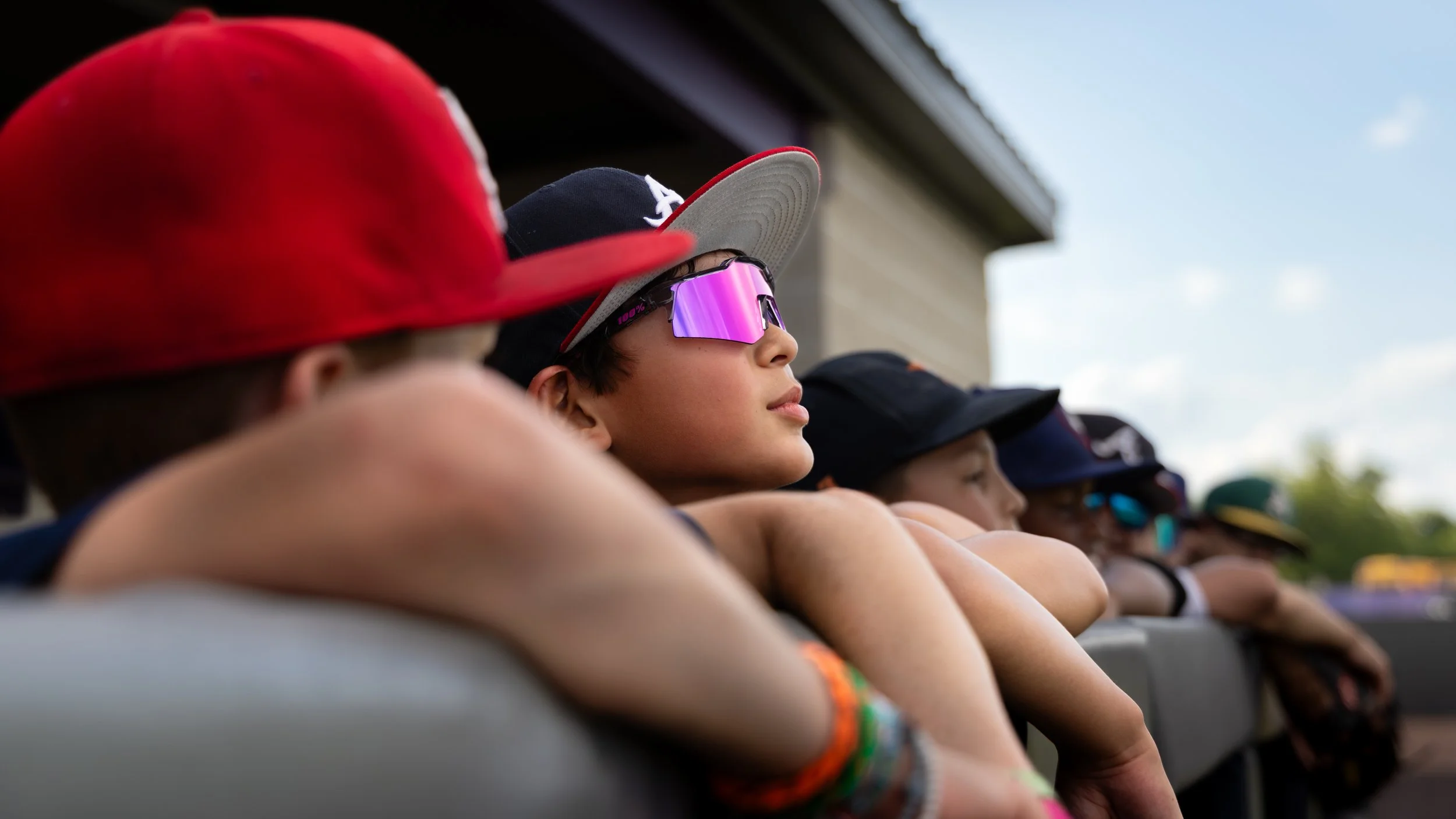 Group of young boys leaning on a railing, wearing caps and sunglasses, watching an outdoor event under a partly cloudy sky.