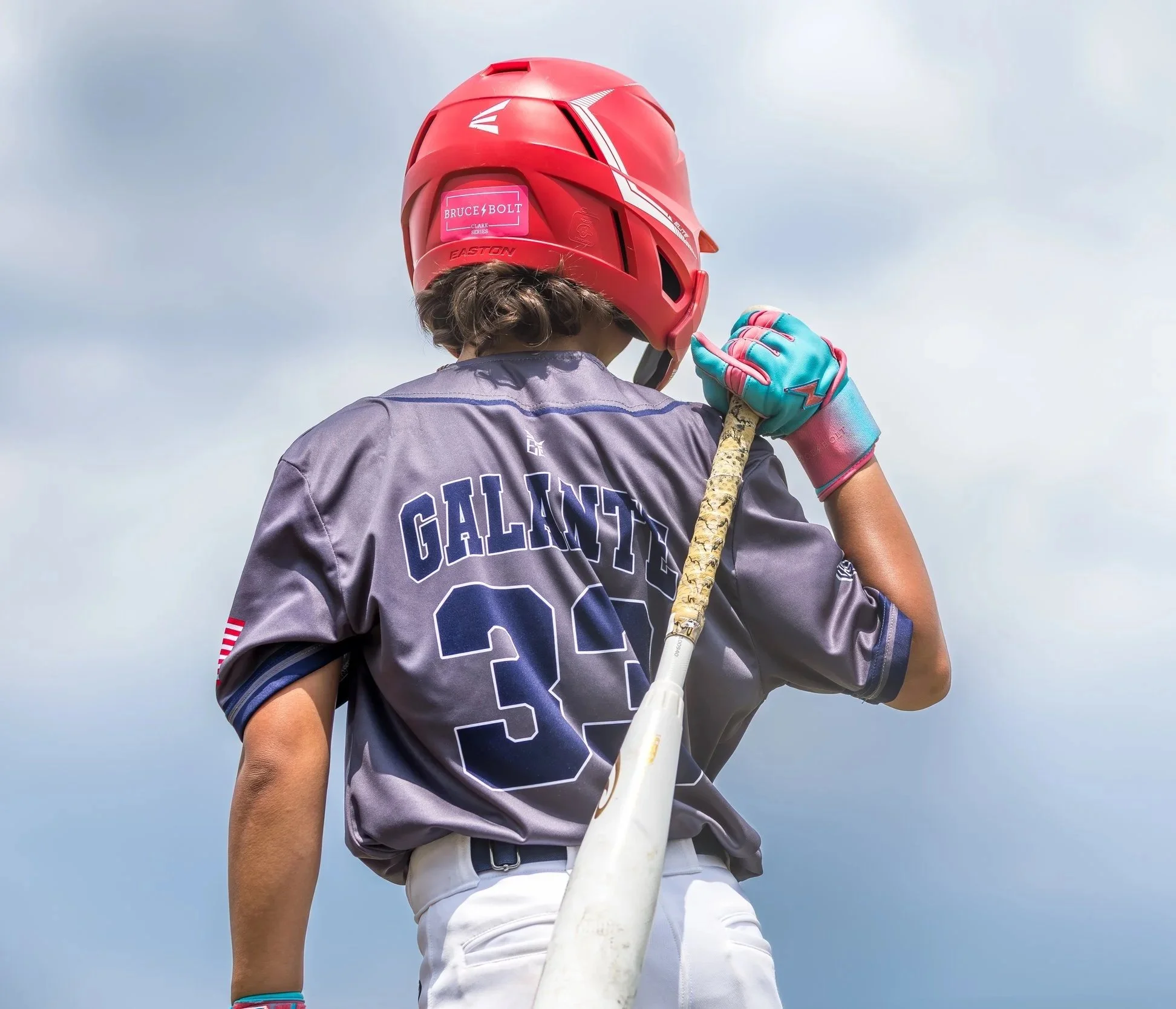 Child wearing a red helmet and a gray sports jersey with the name 'GALLANT' and the number '33' on the back, holding a baseball bat over the shoulder against a cloudy sky.