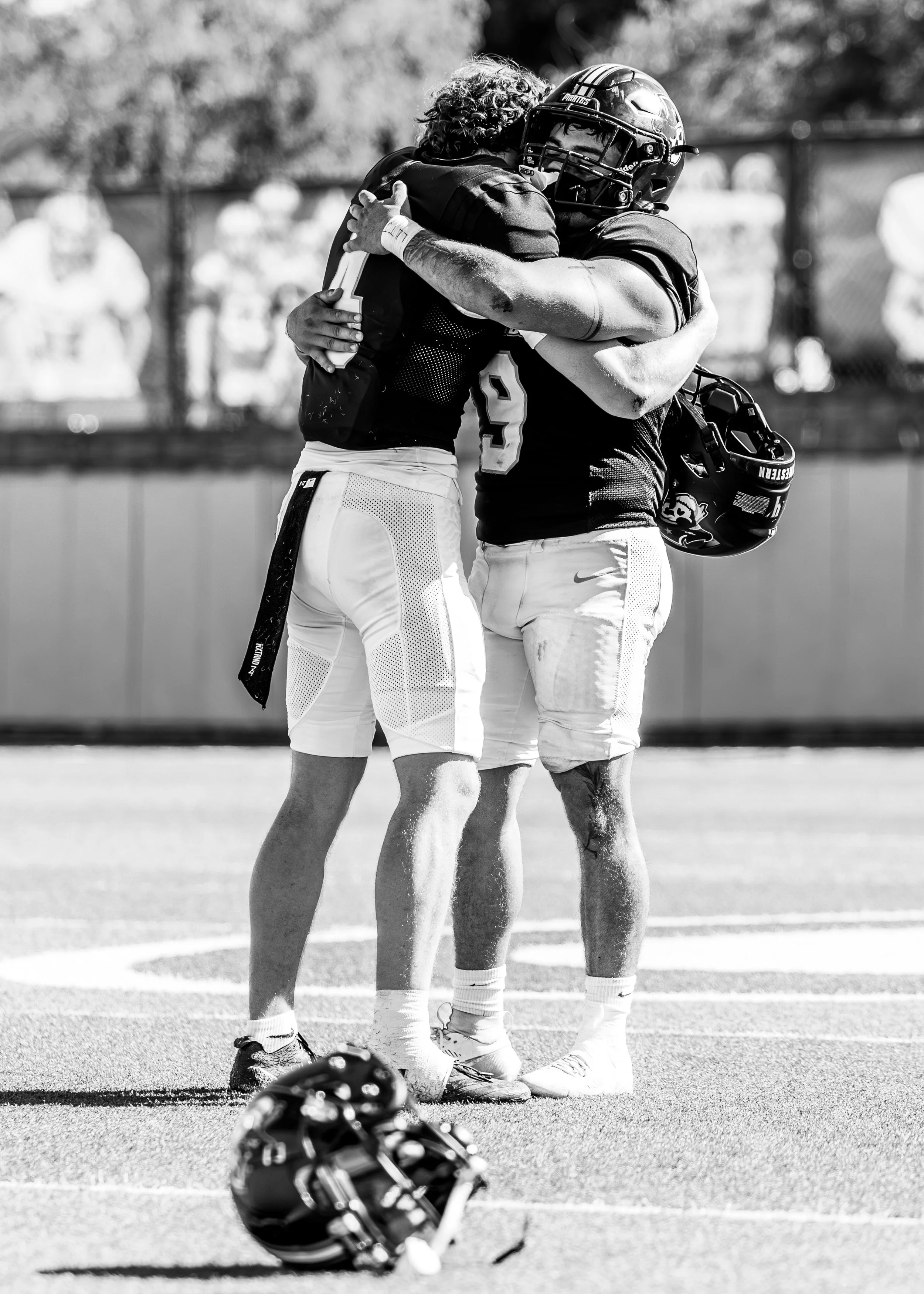 Two football players hugging on the field, one with a helmet in hand and the other wearing a helmet. The field has yard lines, and a third helmet is on the ground nearby.