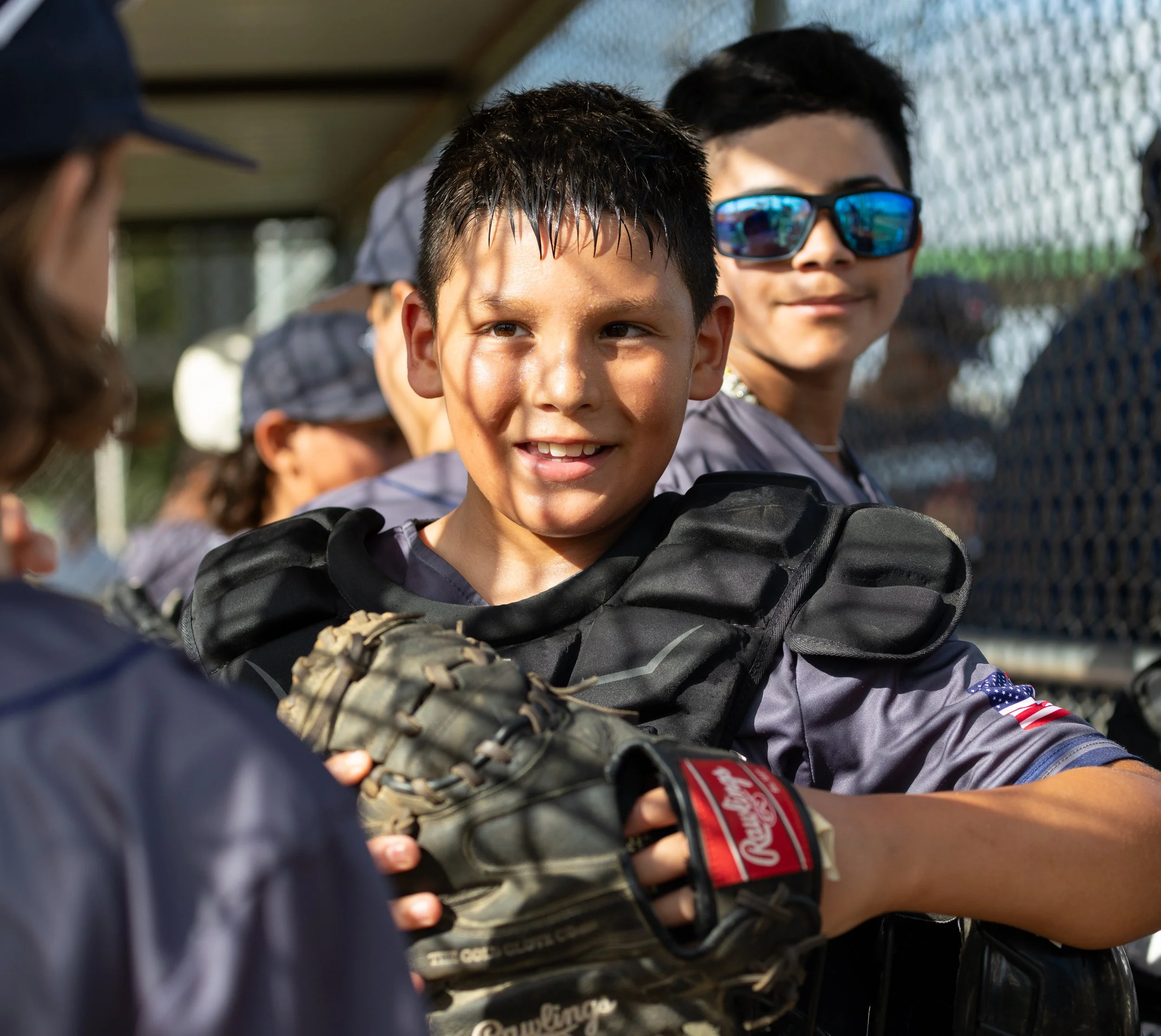 Young baseball players in uniform with gloves on a baseball field, smiling and interacting with each other.