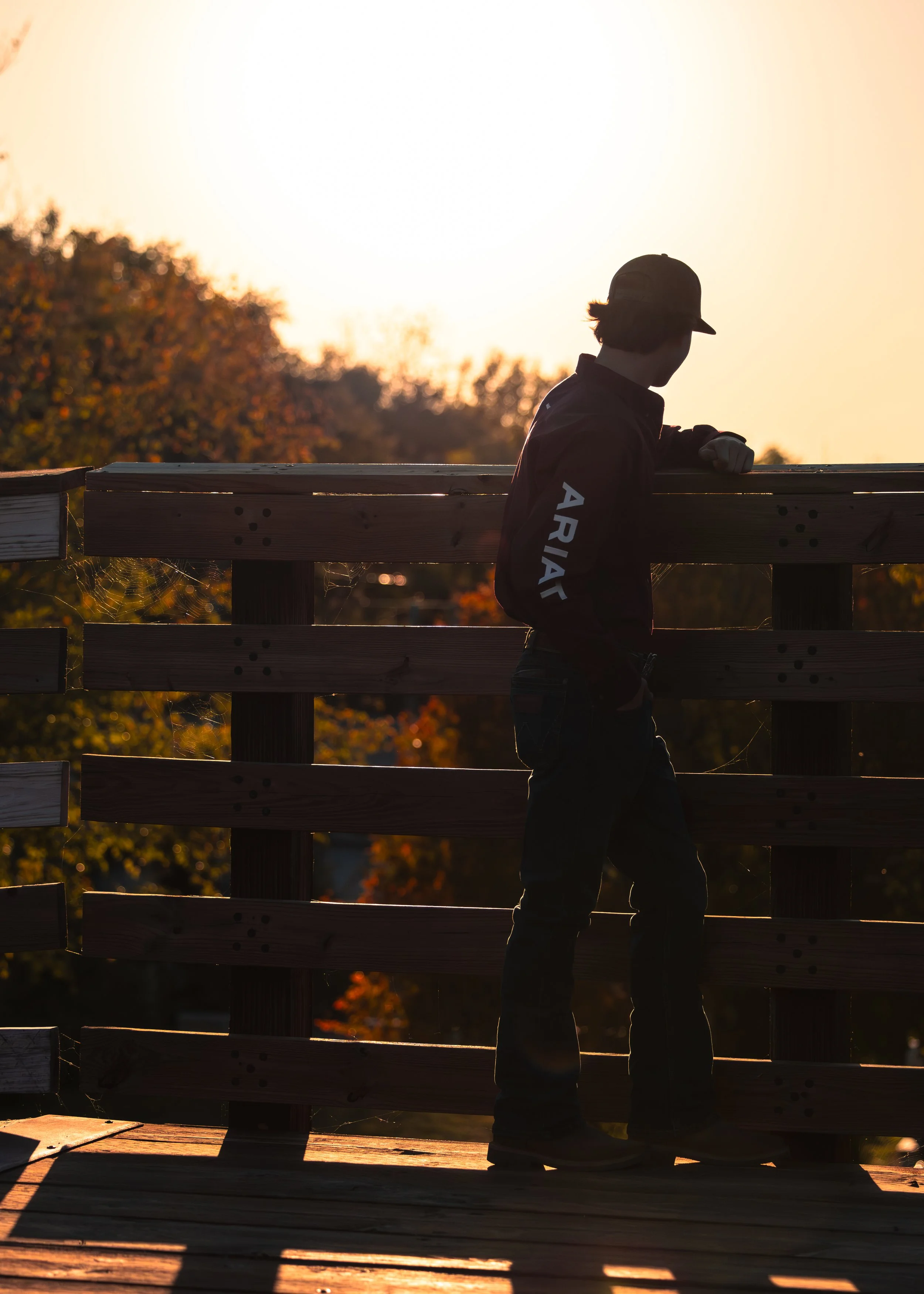 Silhouette of a young boy leaning on a wooden railing at sunset, wearing a cap and jacket with 'ARIAT' on the sleeve, with trees and a clear sky in the background.