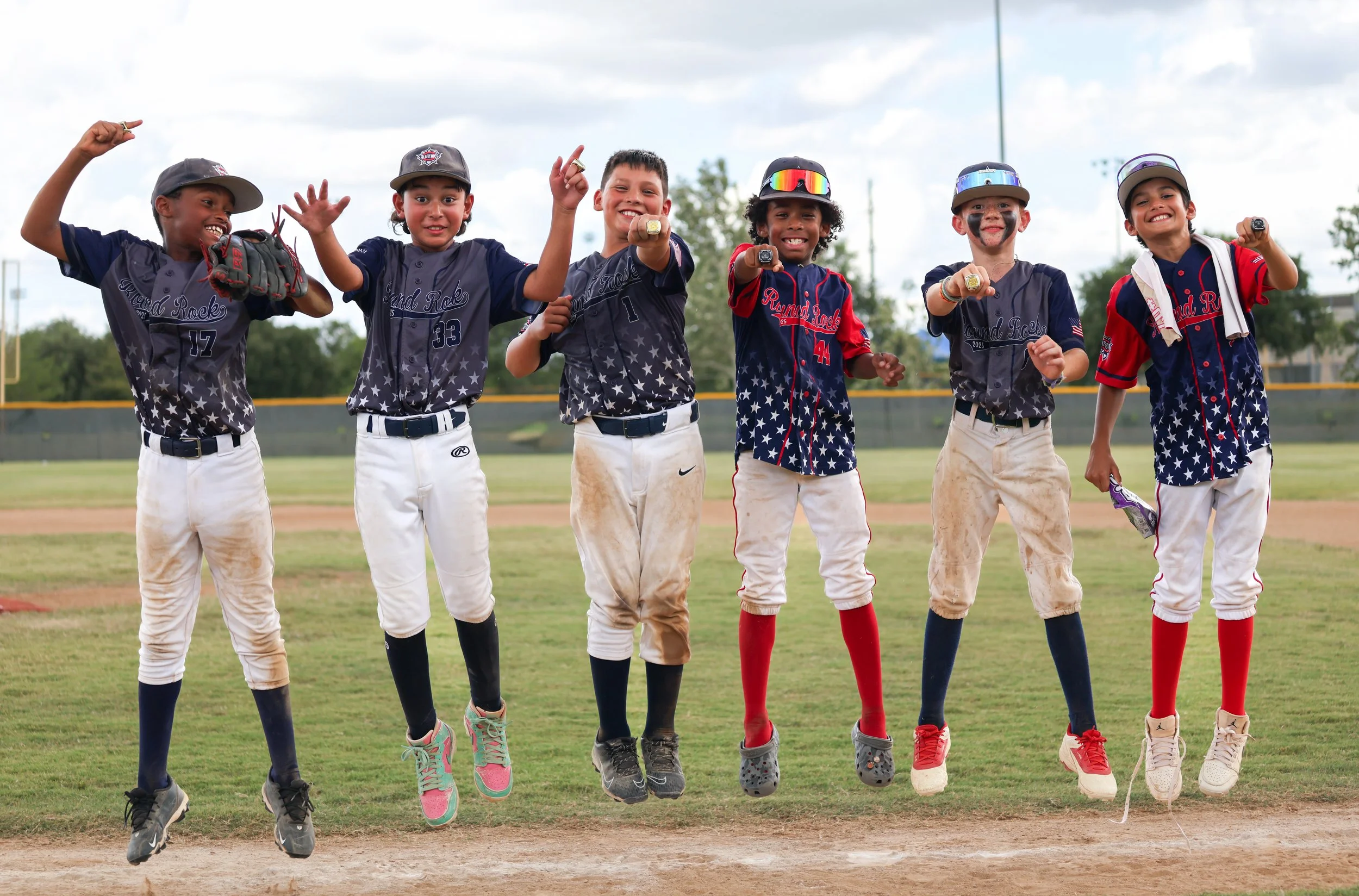 Group of six young boys in baseball uniforms jumping in the air on a baseball field, smiling, during daytime.