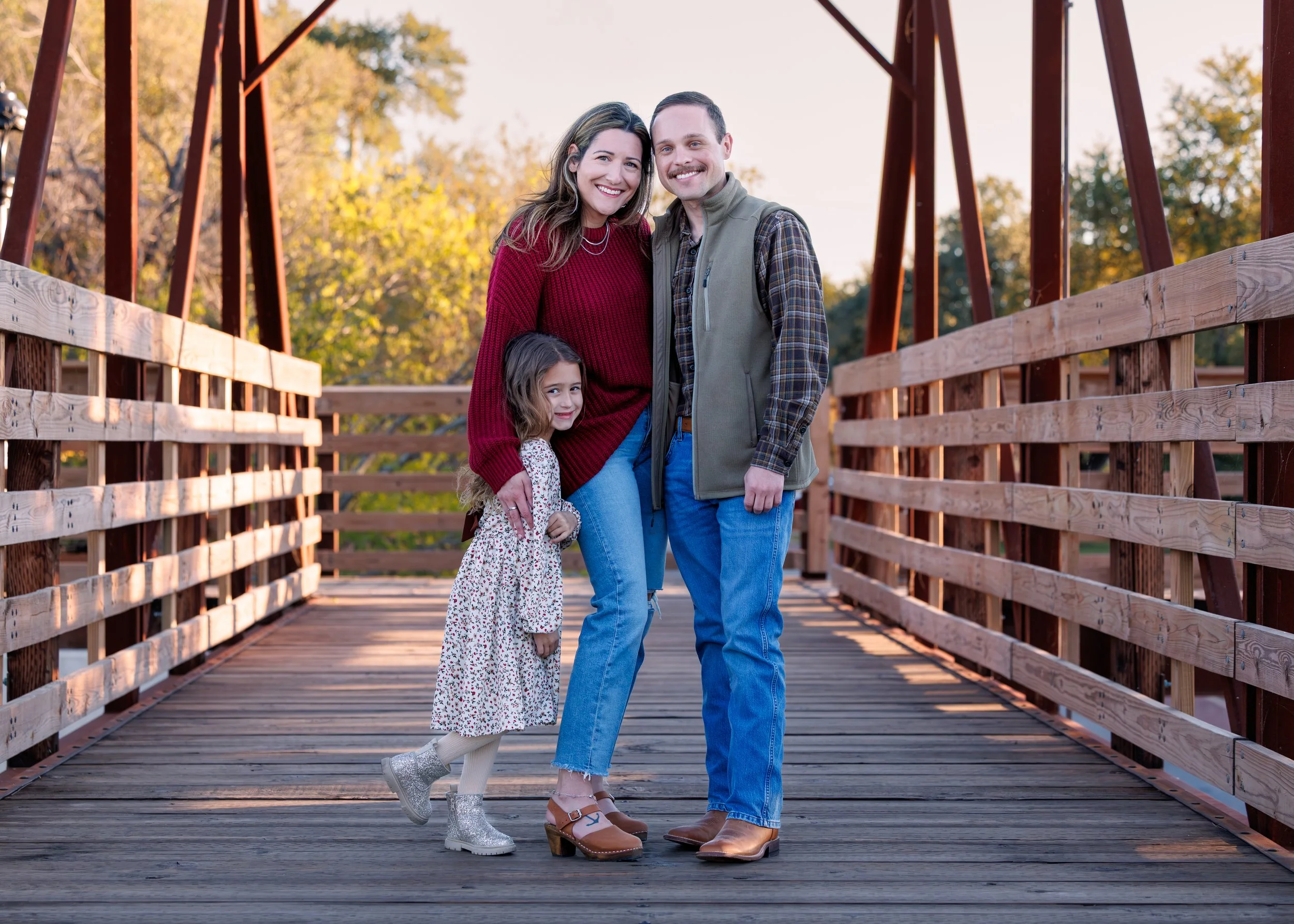 A family of four standing on a wooden bridge outdoors in autumn, smiling at the camera. They include a woman in a red sweater, a man in a plaid shirt and vest, a young girl in a floral dress and boots, and a boy partially visible behind the woman.