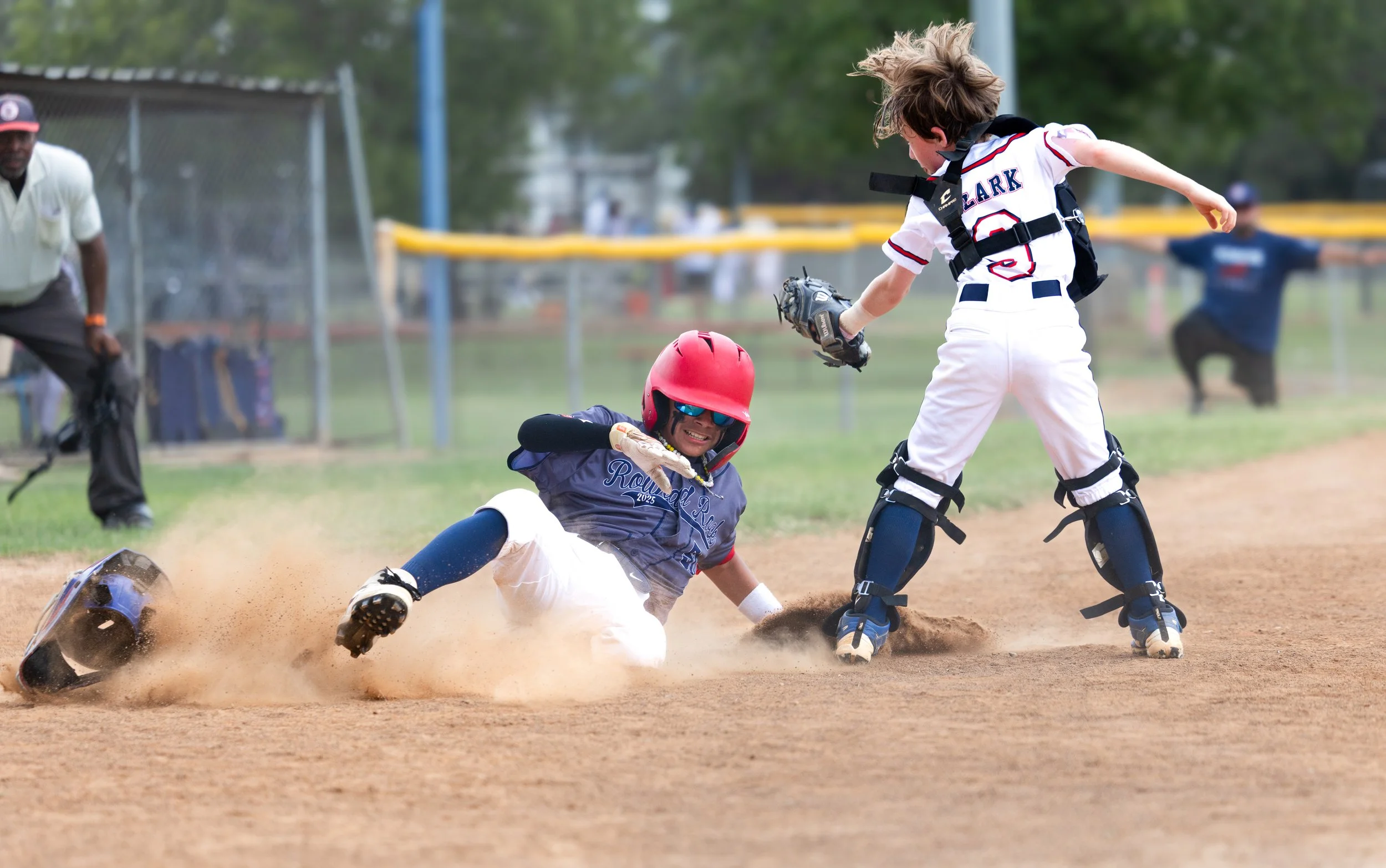A young baseball player sliding into home plate while another player attempts to tag him out, with an umpire and a fielder in the background.