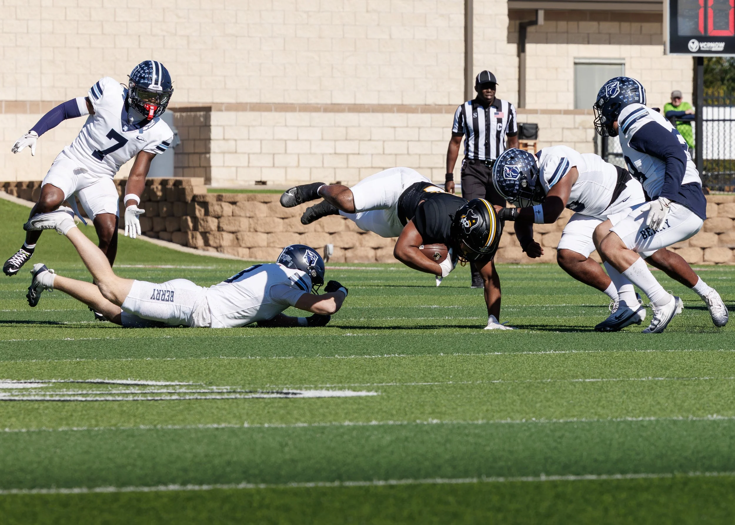 A football game in progress with players from two teams, one team in white and blue uniforms and the other in black uniforms, on a green field. One player in black is holding a football and diving forward while being tackled by players in white. An o