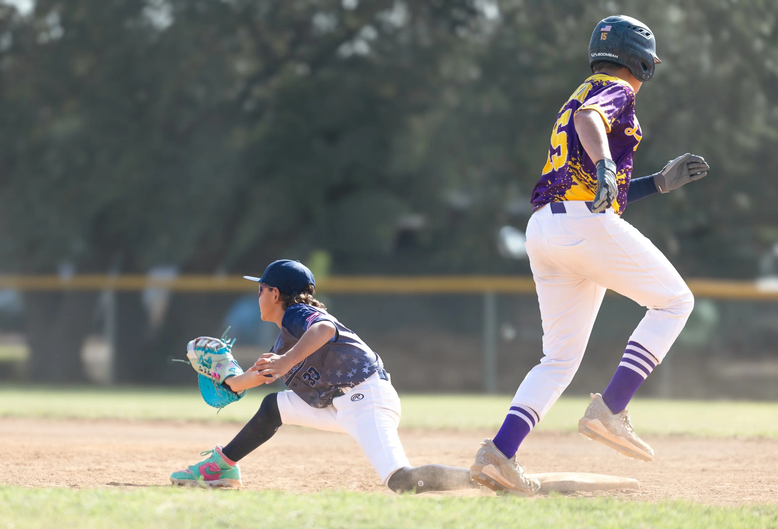 A young baseball player in a purple and yellow uniform running past a teammate in a dark uniform and a cap, who is sliding into a base on a baseball field.