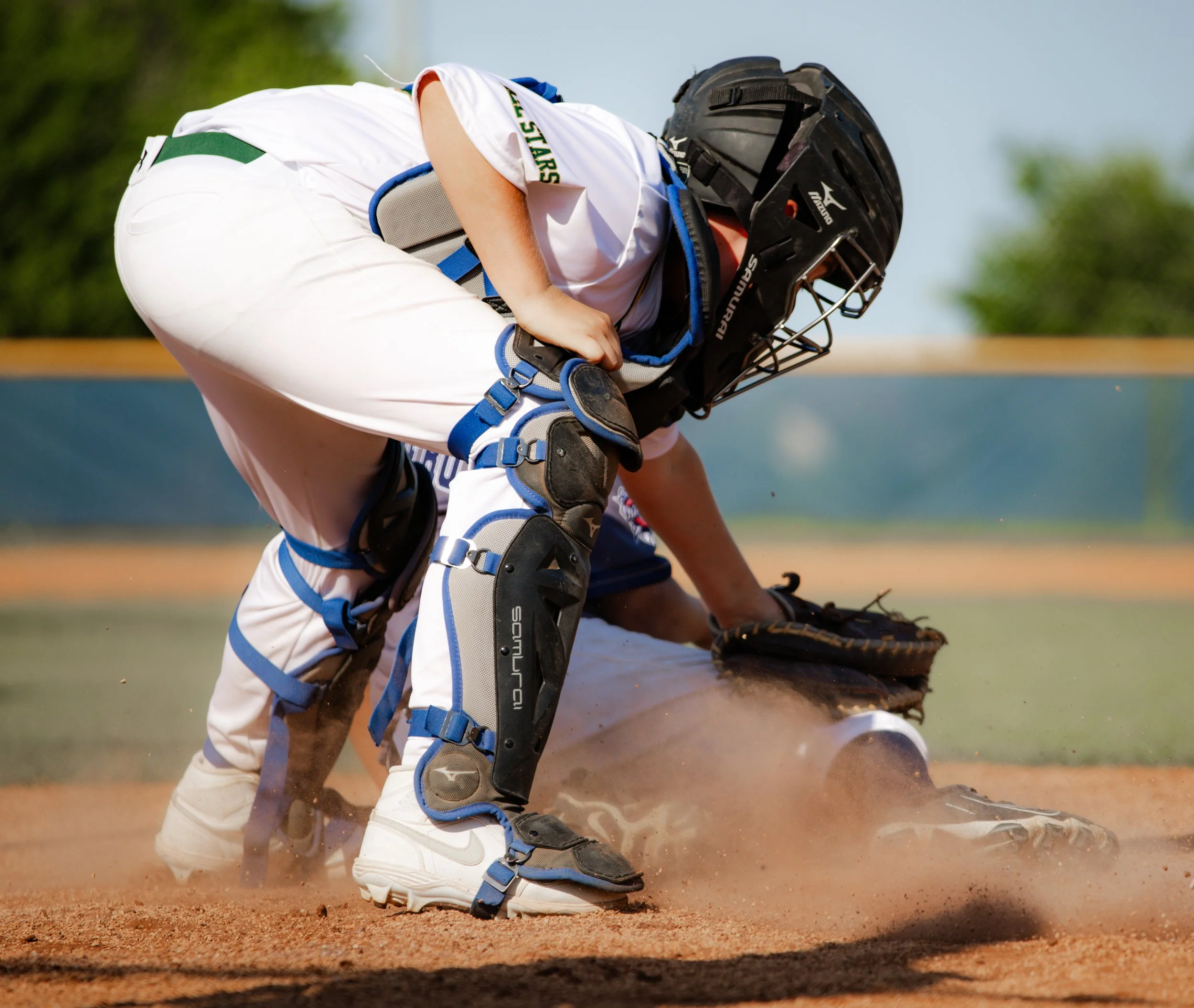 A baseball catcher is crouched behind home plate on the field, wearing a helmet, chest protector, shin guards, and mitt, ready to catch a pitch during a game.