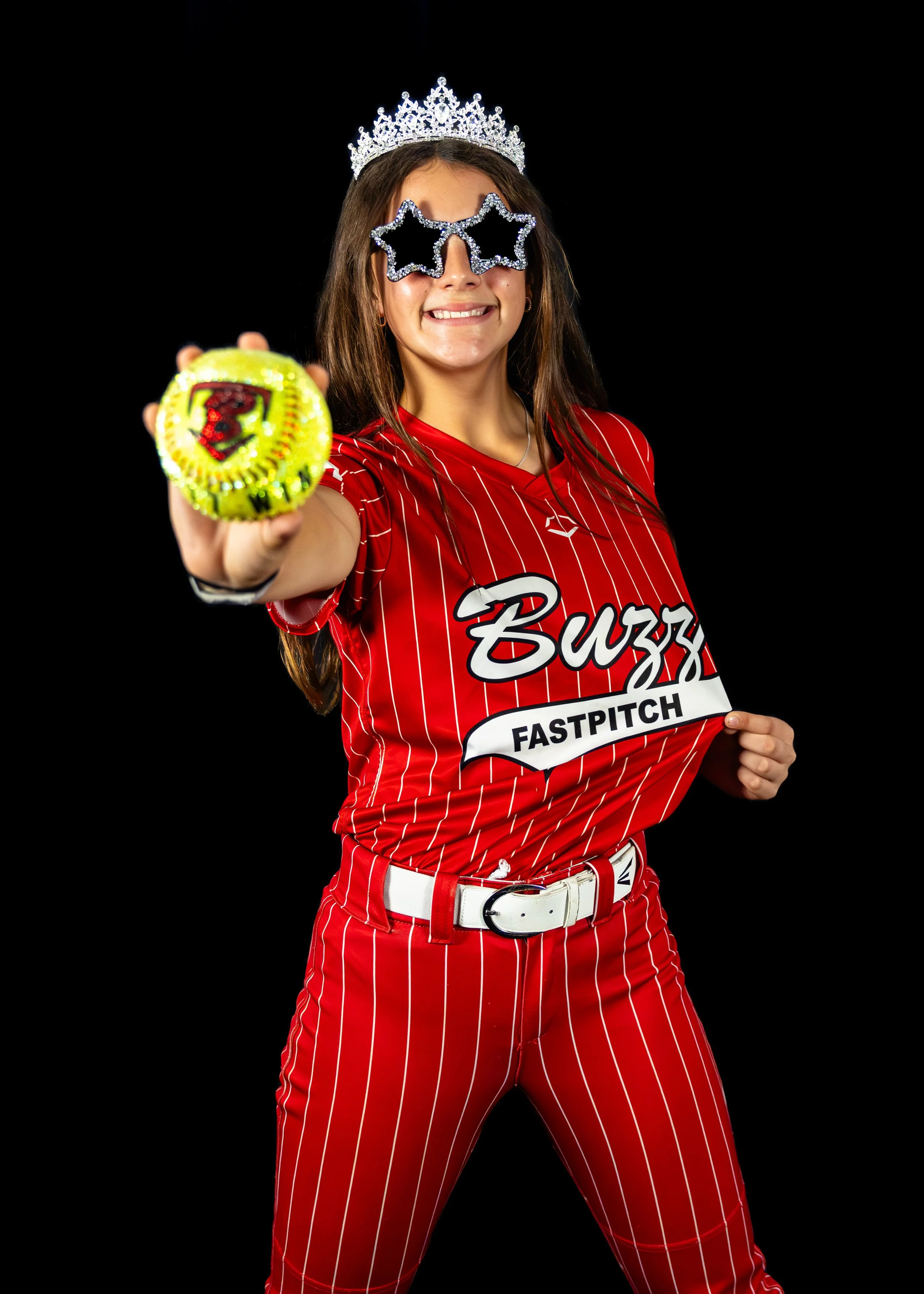 Woman wearing a red sports jersey and striped pants, holding a badge, with star-shaped glasses and a crown, smiling on a black background.