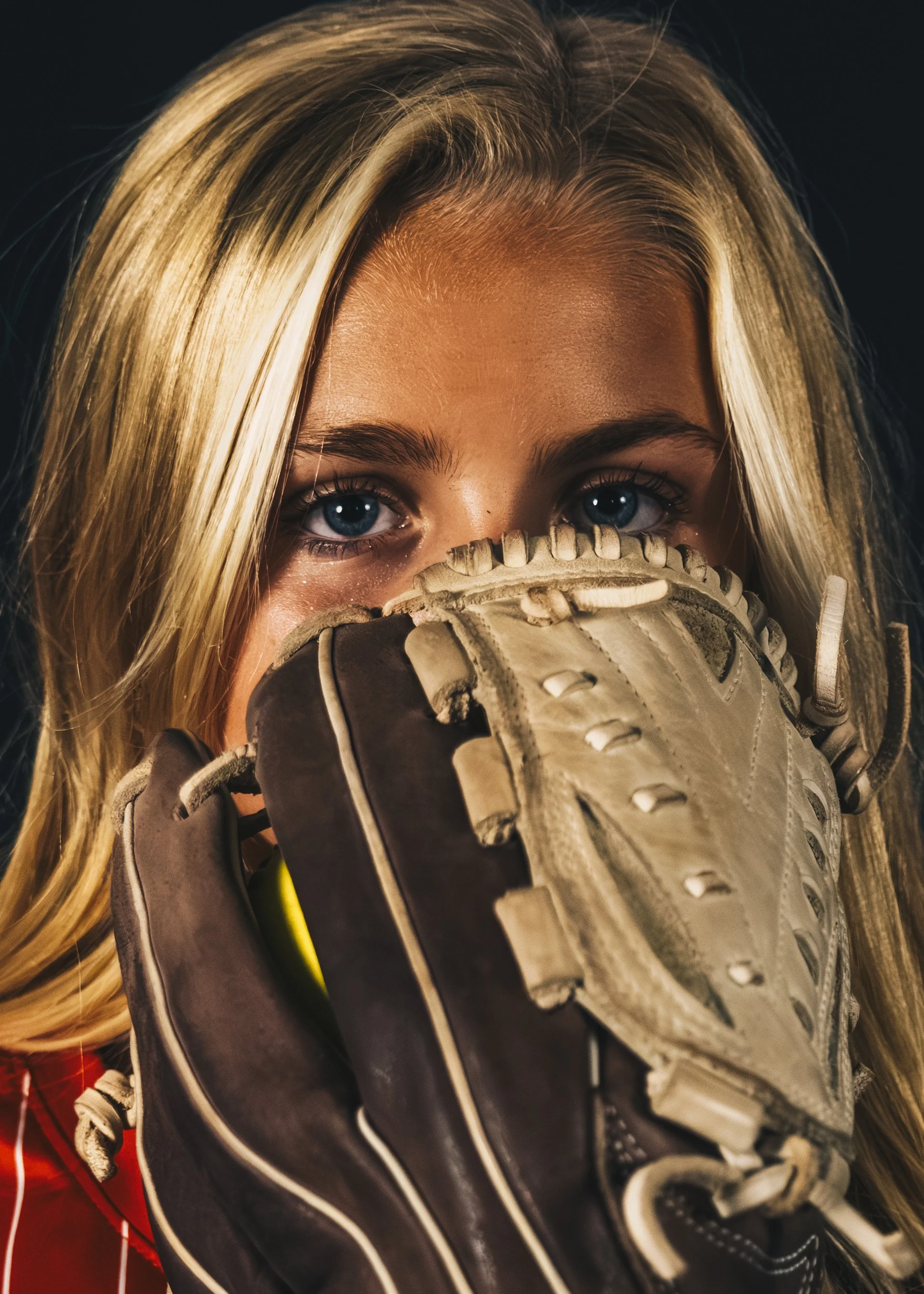 Close-up of a young woman with blonde hair and blue eyes holding a baseball glove near her face.