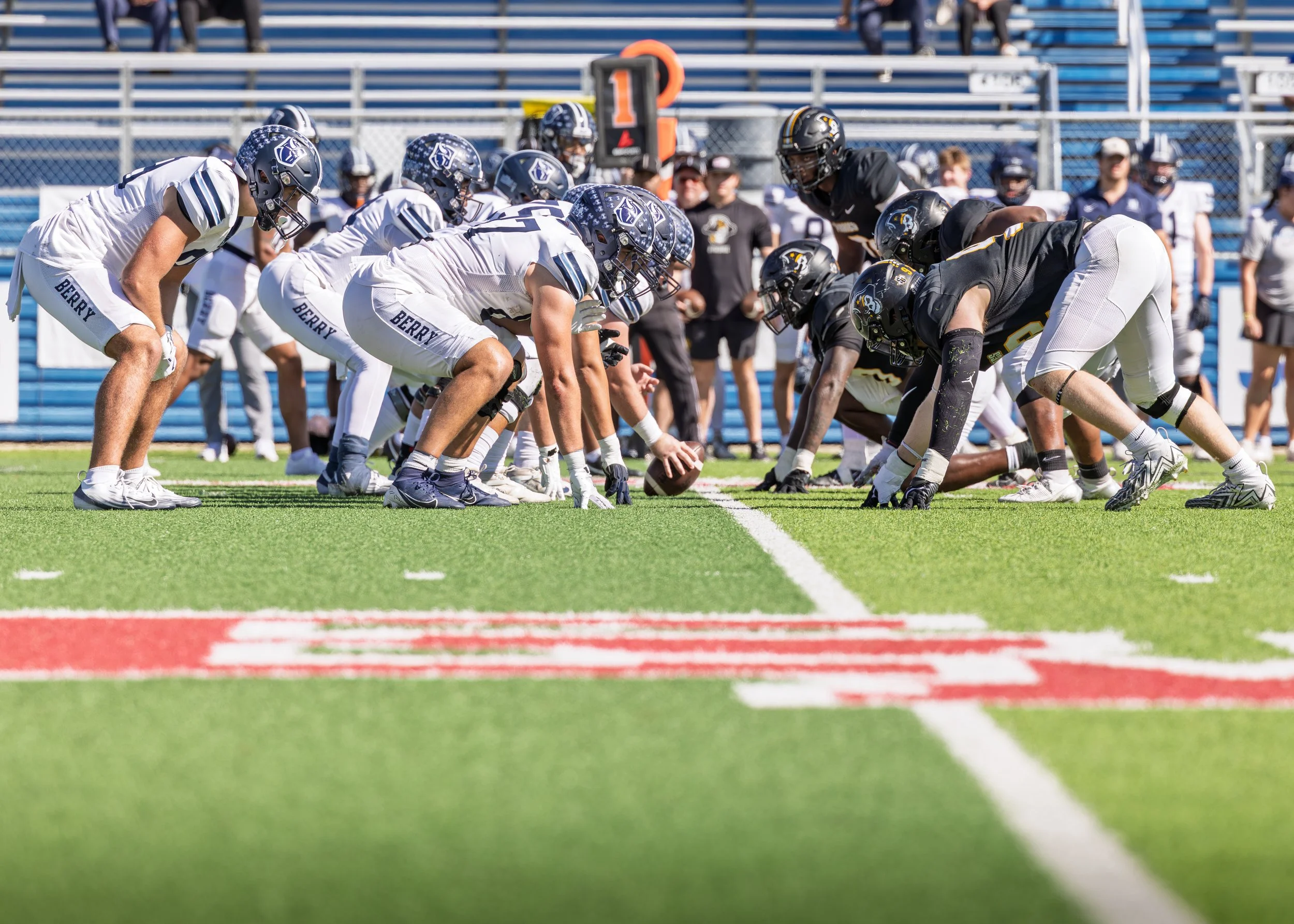 American football players in a line of scrimmage on a football field preparing for the snap, with players in white and black uniforms and coaches and spectators in the background.