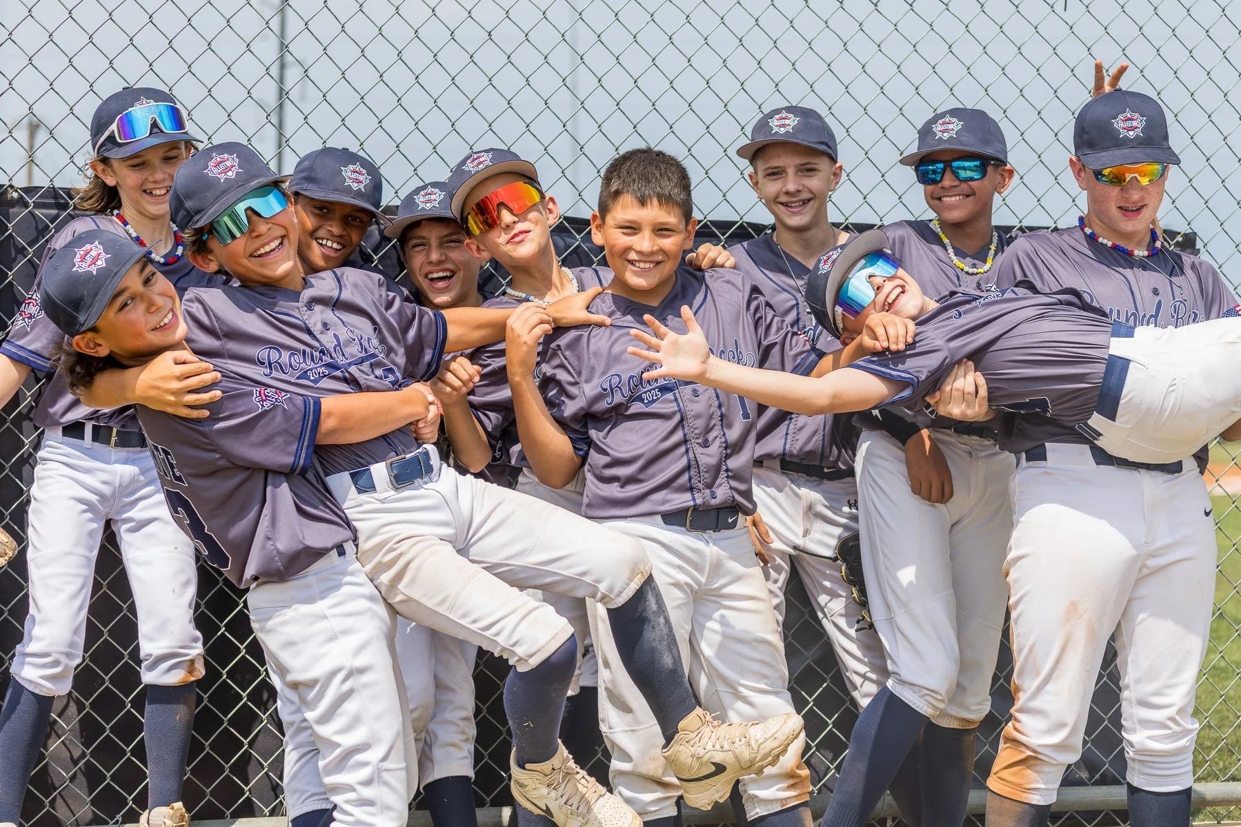 Group of young boys in gray baseball uniforms celebrating after a game, some wearing sunglasses, standing in front of a chain-link fence.