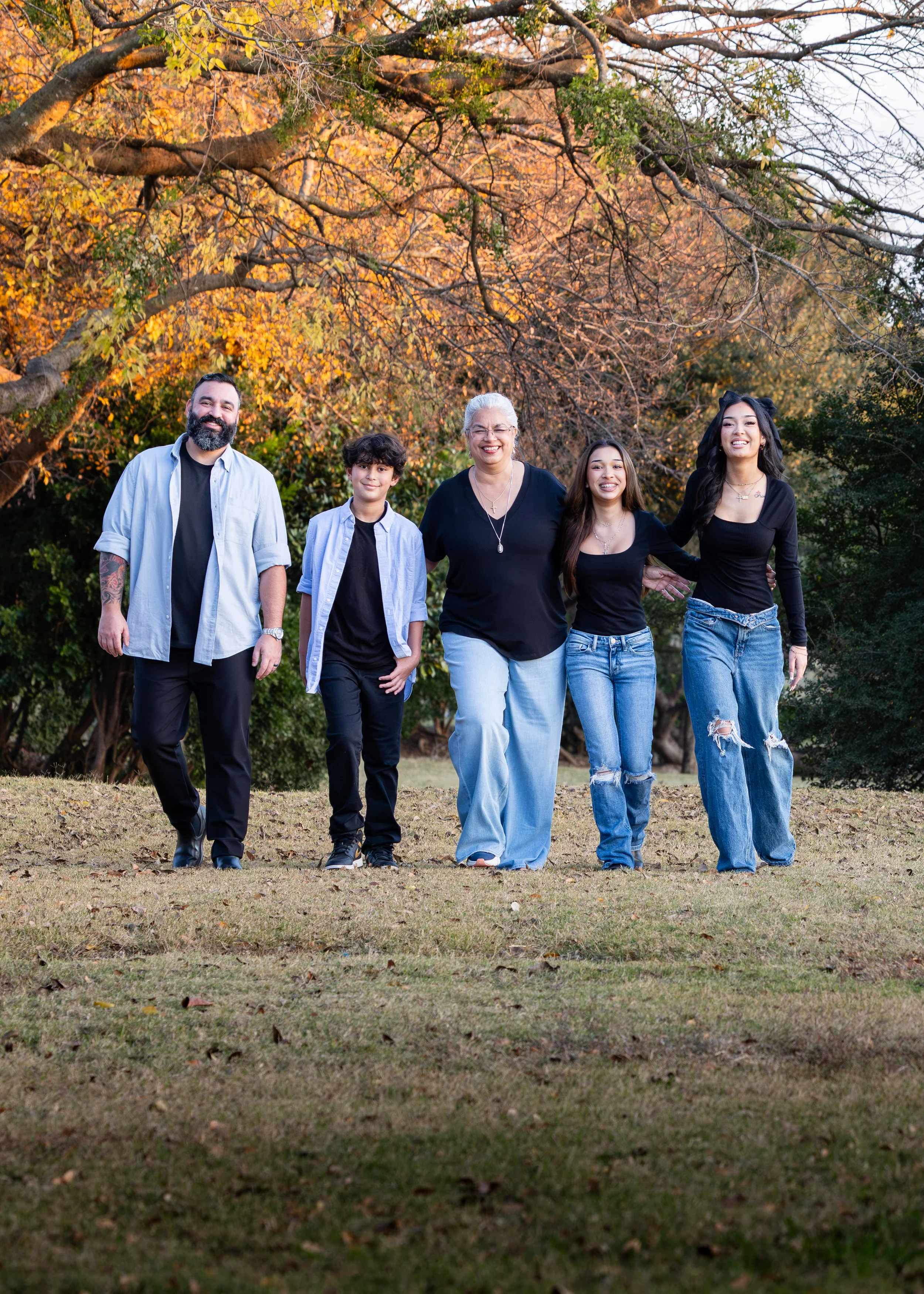 A group of five people, including a man, a boy, an older woman, and two young women, walking outdoors on grass during autumn in a park with trees showing fall colors in the background.