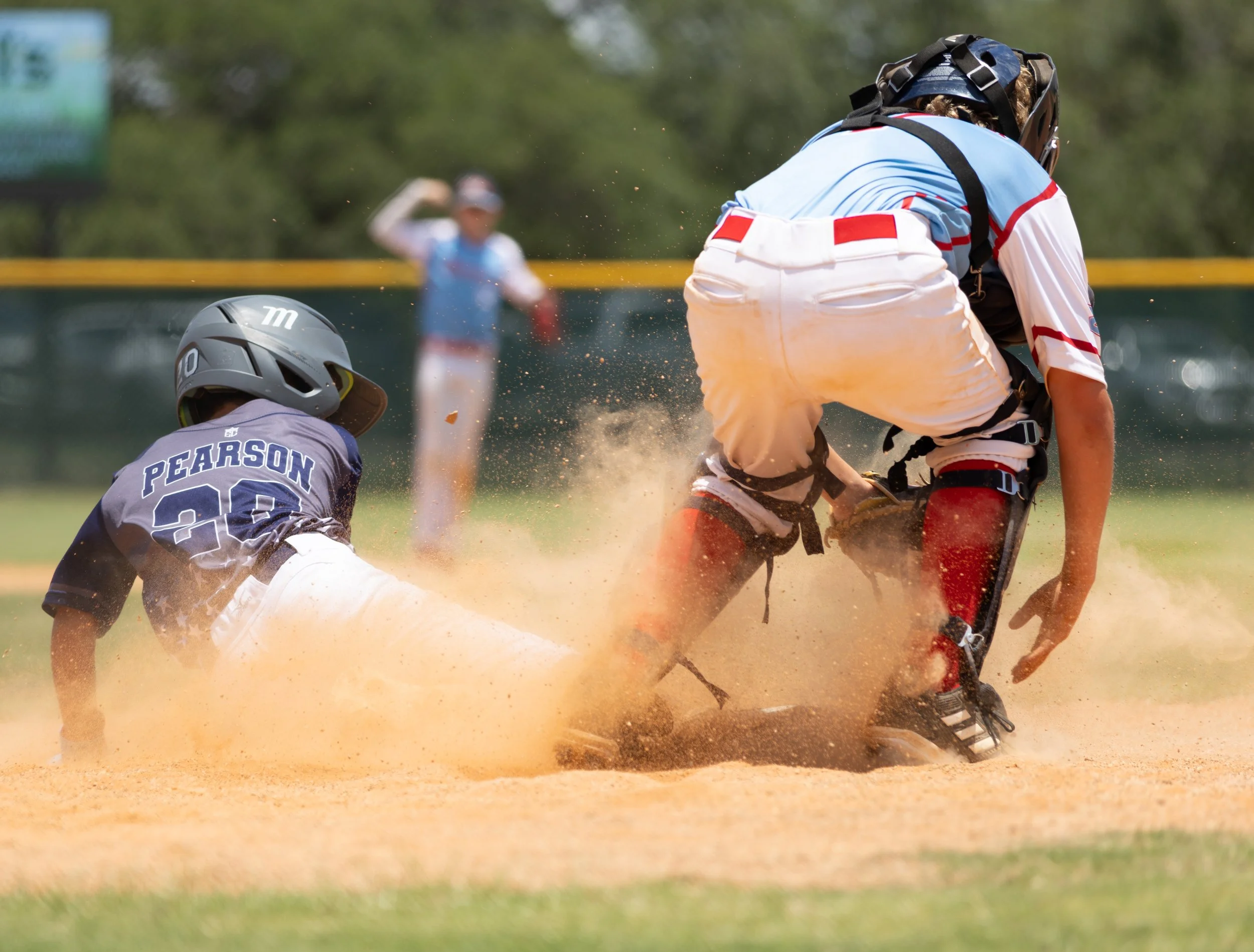 Two baseball players, one in a gray uniform labeled 'PEARSON' and the other in a white and red uniform, are competing for the base on a dirt field. The player in gray is sliding headfirst, while the player in white attempts to tag him. A blurry figur