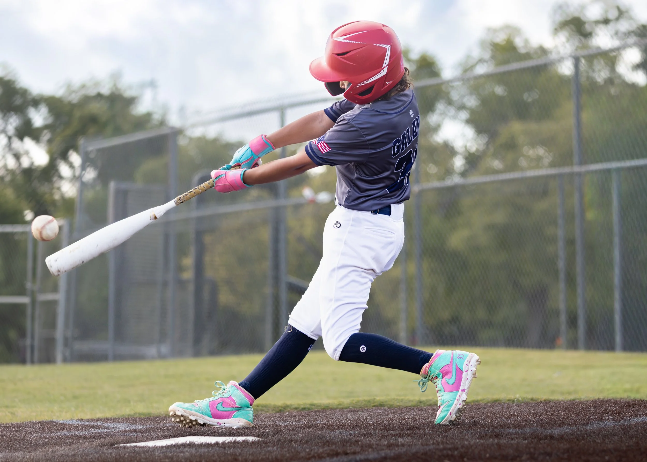 A young girl in a baseball uniform and helmet swings a bat to hit a baseball on a field.