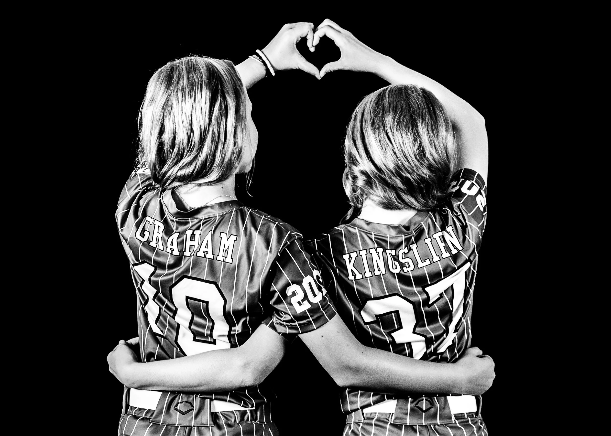 Two female athletes in striped sports jerseys with their names, Graham and Kingslien, and numbers 8 and 3, making a heart shape with their hands.