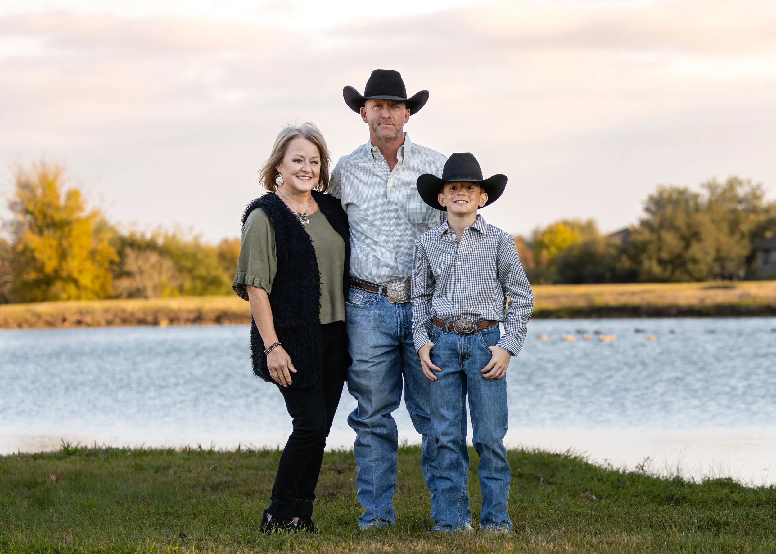 A family of three wearing cowboy hats standing outdoors near a water body during fall, with trees showing autumn colors in the background.