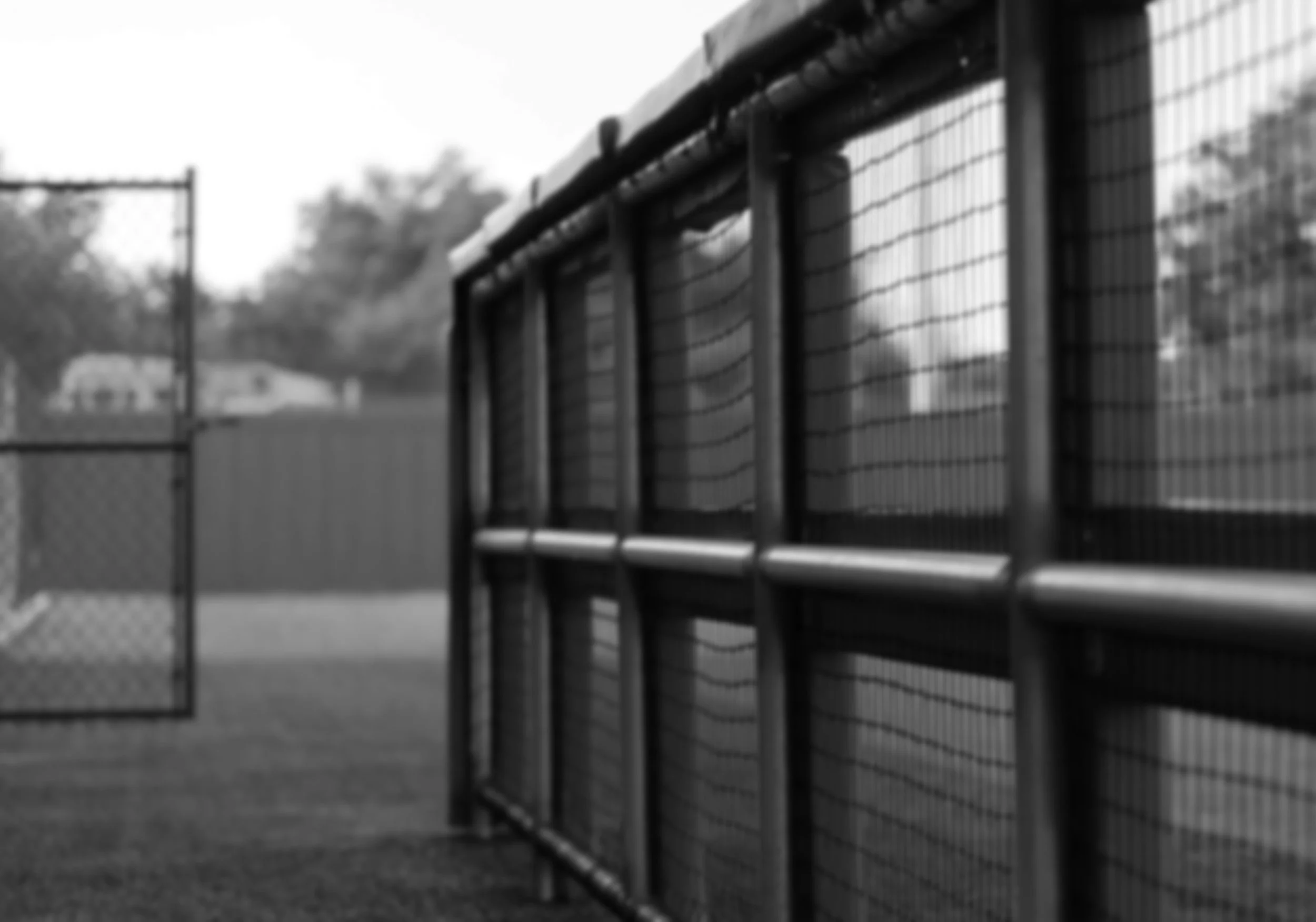 Black and white photo of a chain-link fence surrounding a sports field, with a gate slightly open and trees in the background.