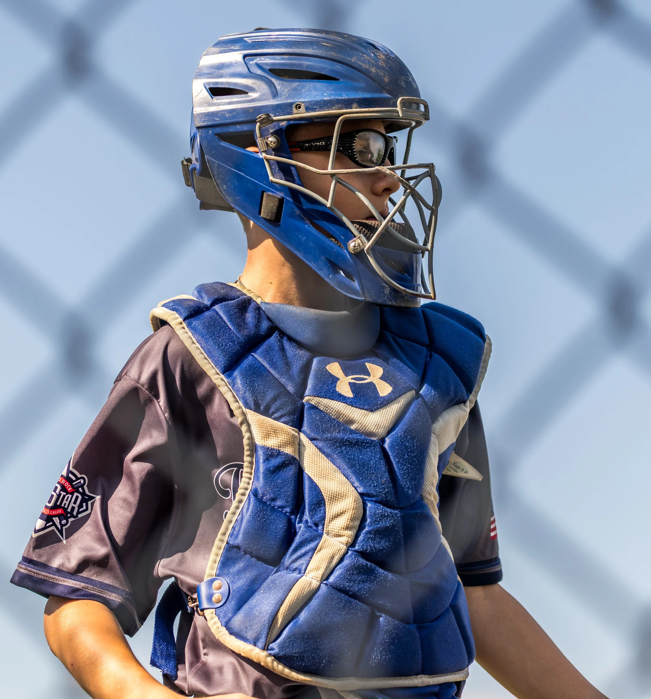 A baseball catcher in uniform, wearing a blue helmet and protective gear, standing behind a chain-link fence.