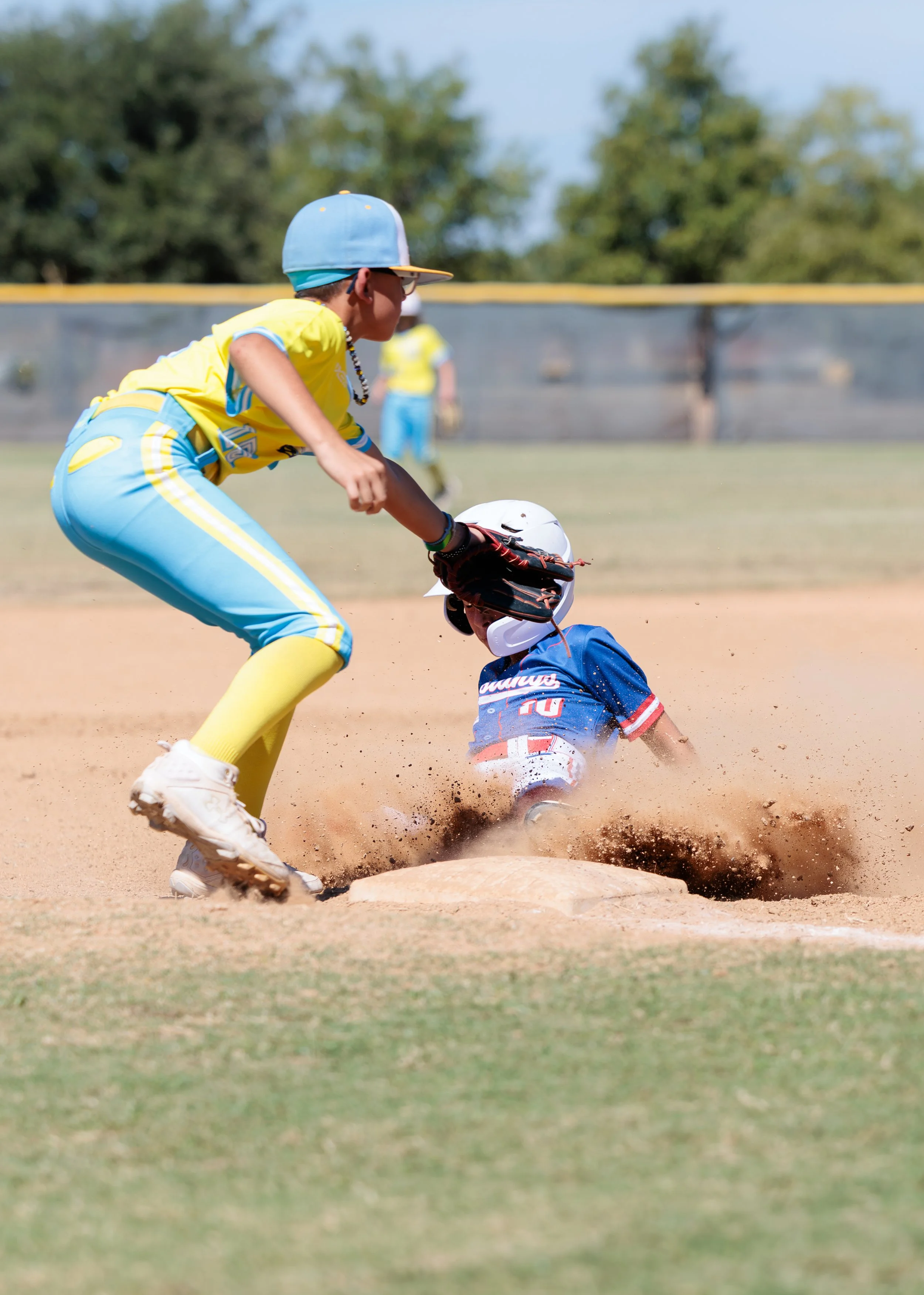 A young baseball player in a yellow and blue uniform tags a sliding player in a blue jersey on third base, dust and dirt flying.