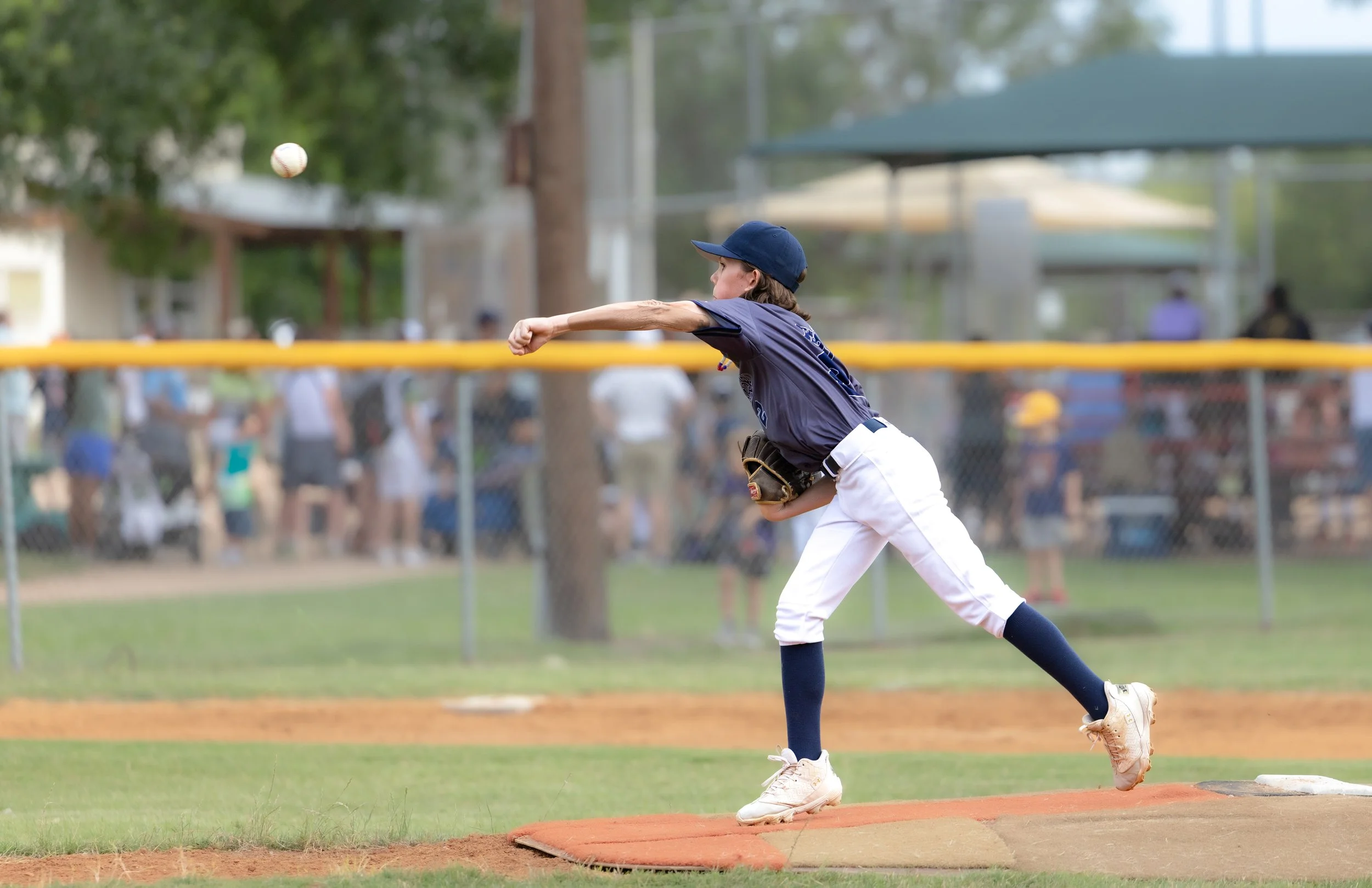 A young baseball player in a navy uniform and cap throws a baseball during a game at a baseball field.