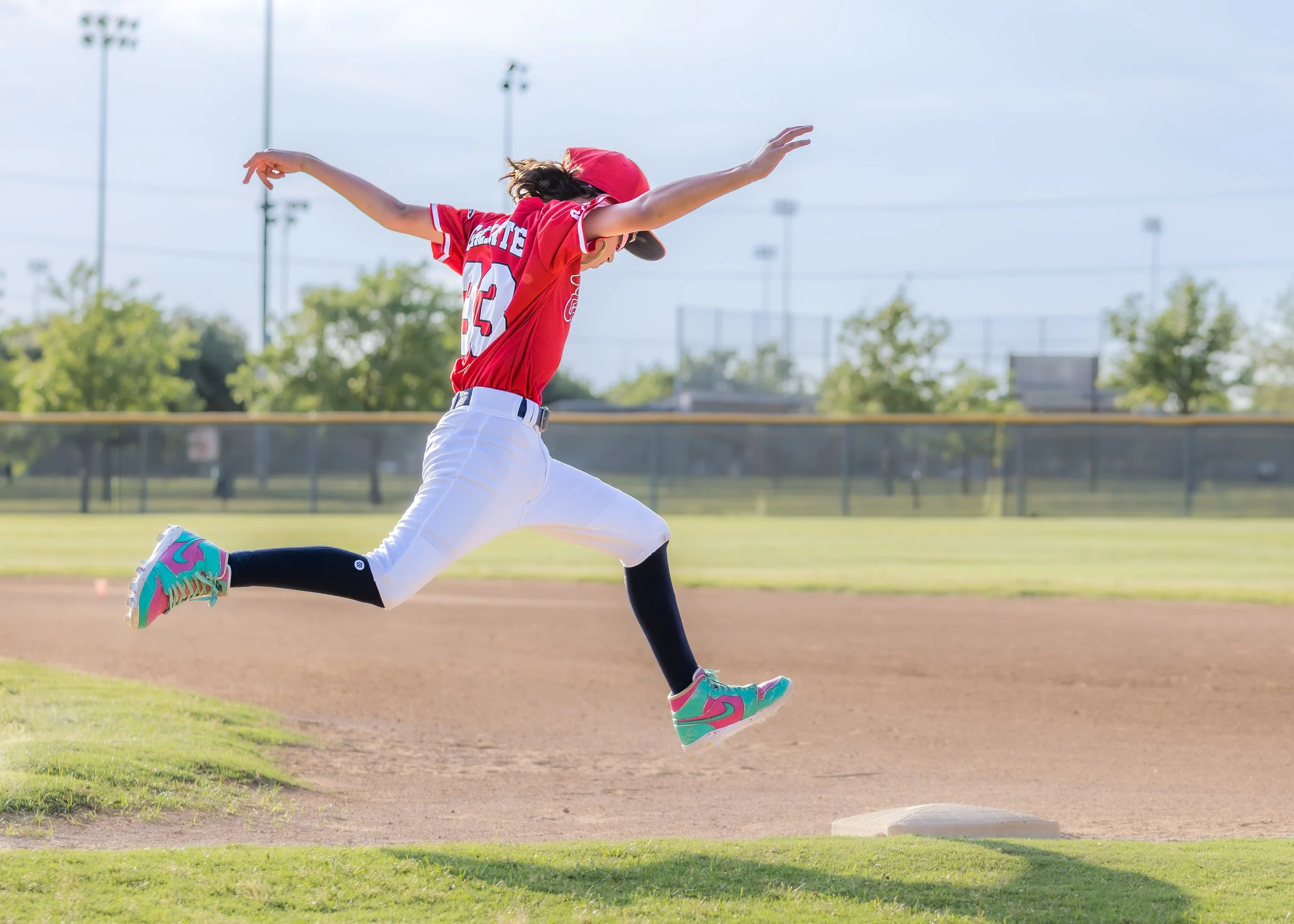 A young girl in a red baseball uniform is running on a baseball field, about to reach a base. She is wearing a red cap, colorful sneakers, and black leggings.