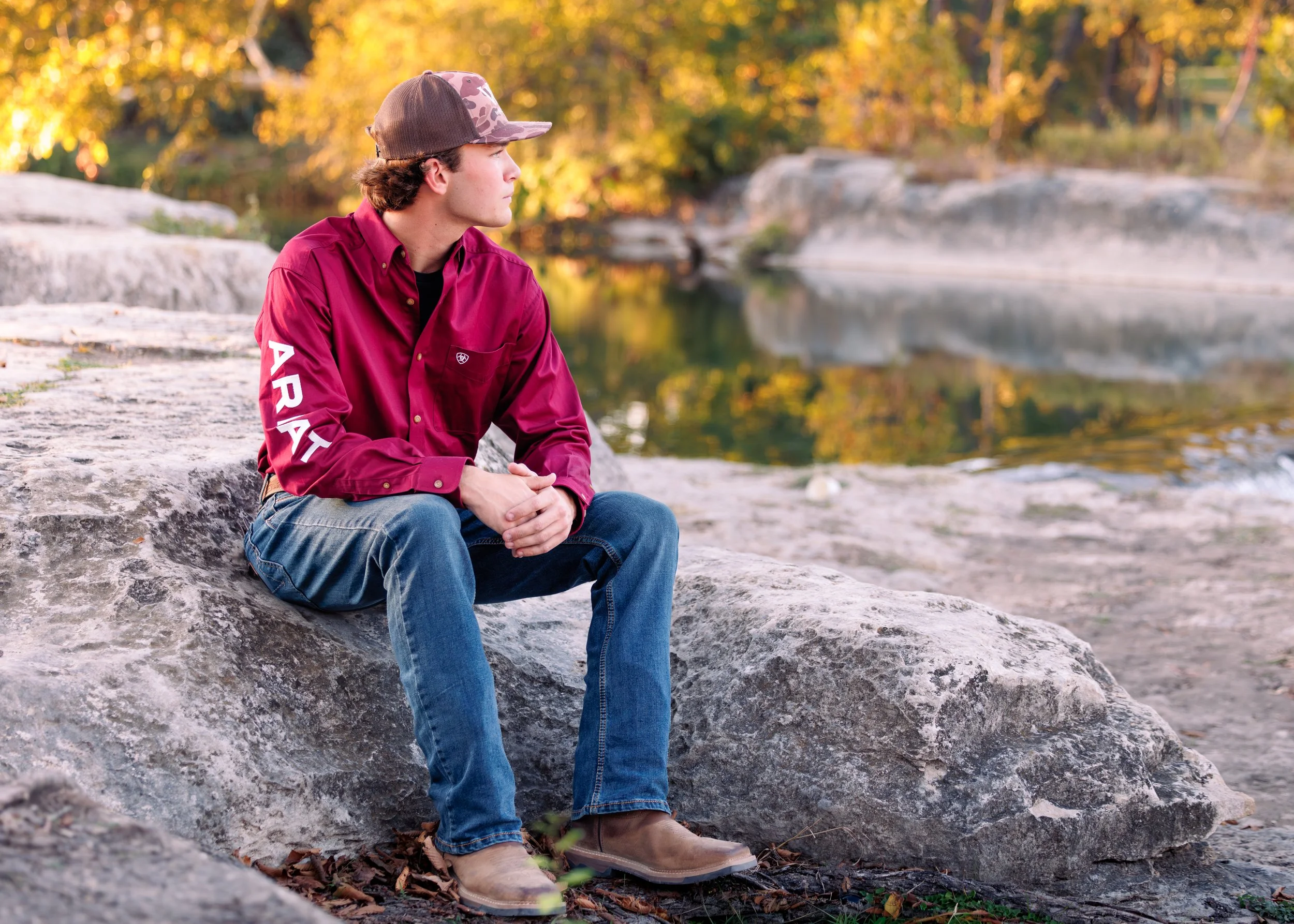 A young man in a red shirt with the word "ARAT" on the sleeve, blue jeans, and a brown cap, sitting on a large rock by a river in an autumn setting, looking to the side.
