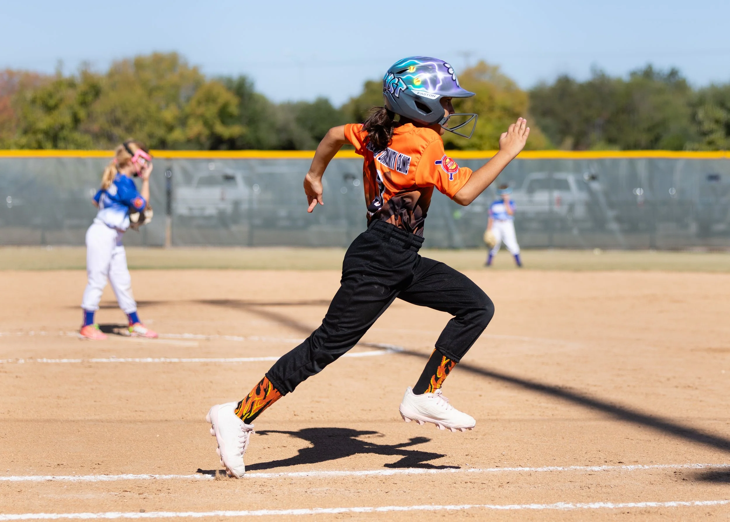 A young girl in a colorful sports uniform running on a baseball field during a game, with other children in the background.
