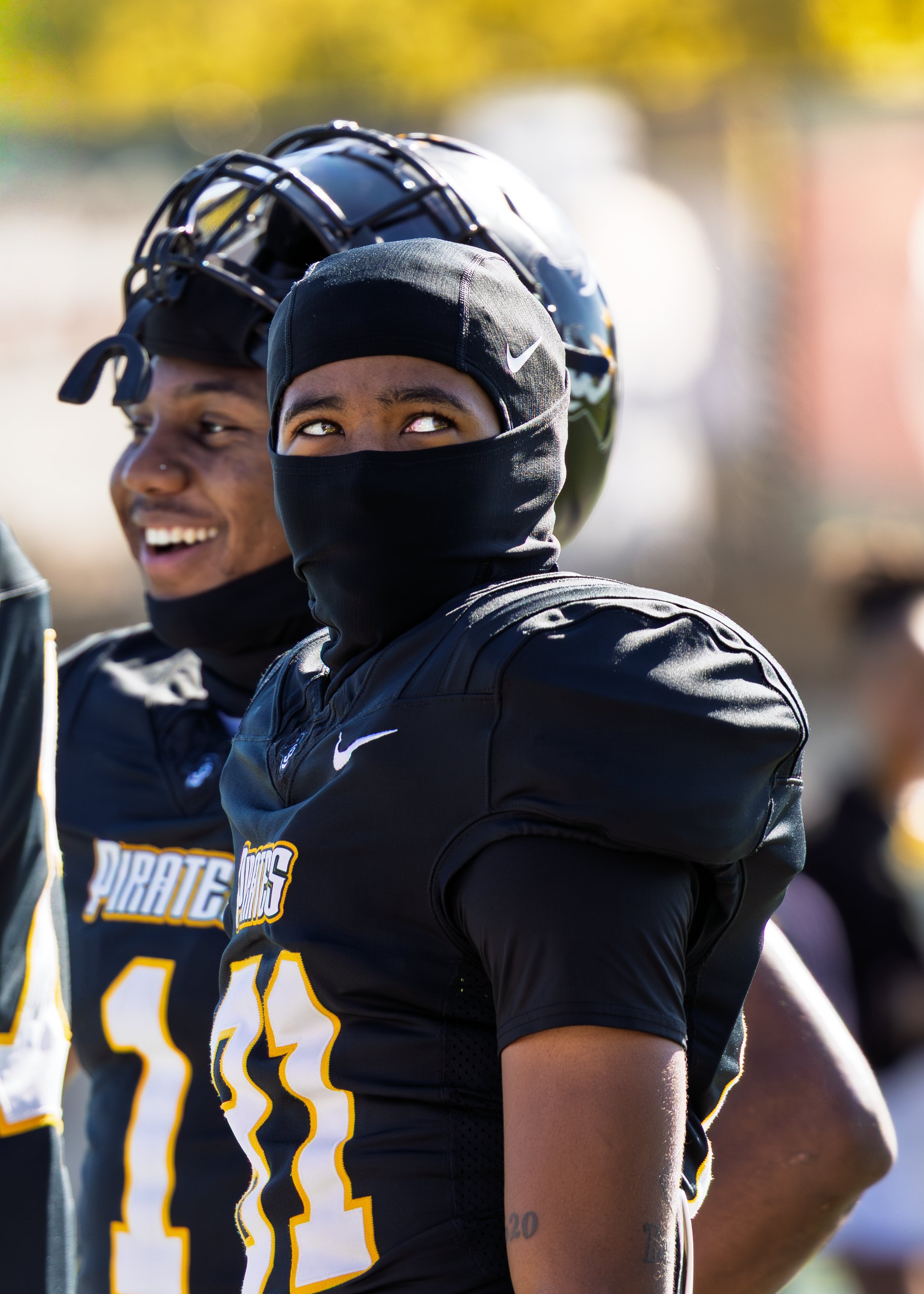 Two football players in black uniforms with yellow and white accents, one wearing a helmet and the other with a black face covering, standing outdoors under sunlight.