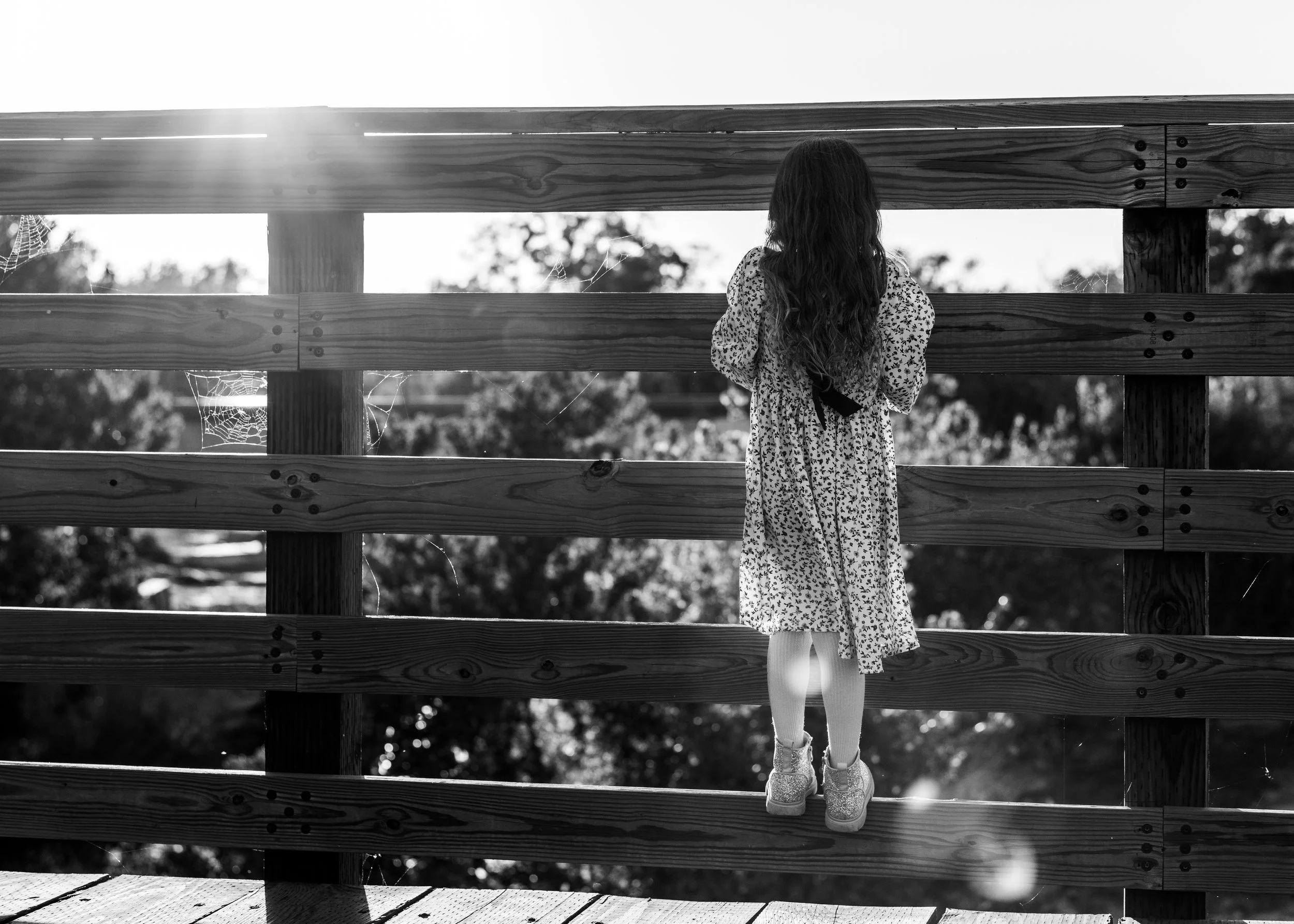 Young girl with long hair in a floral dress and sneakers stands on a wooden bridge, looking out beyond the railing with sunlight shining in the background.