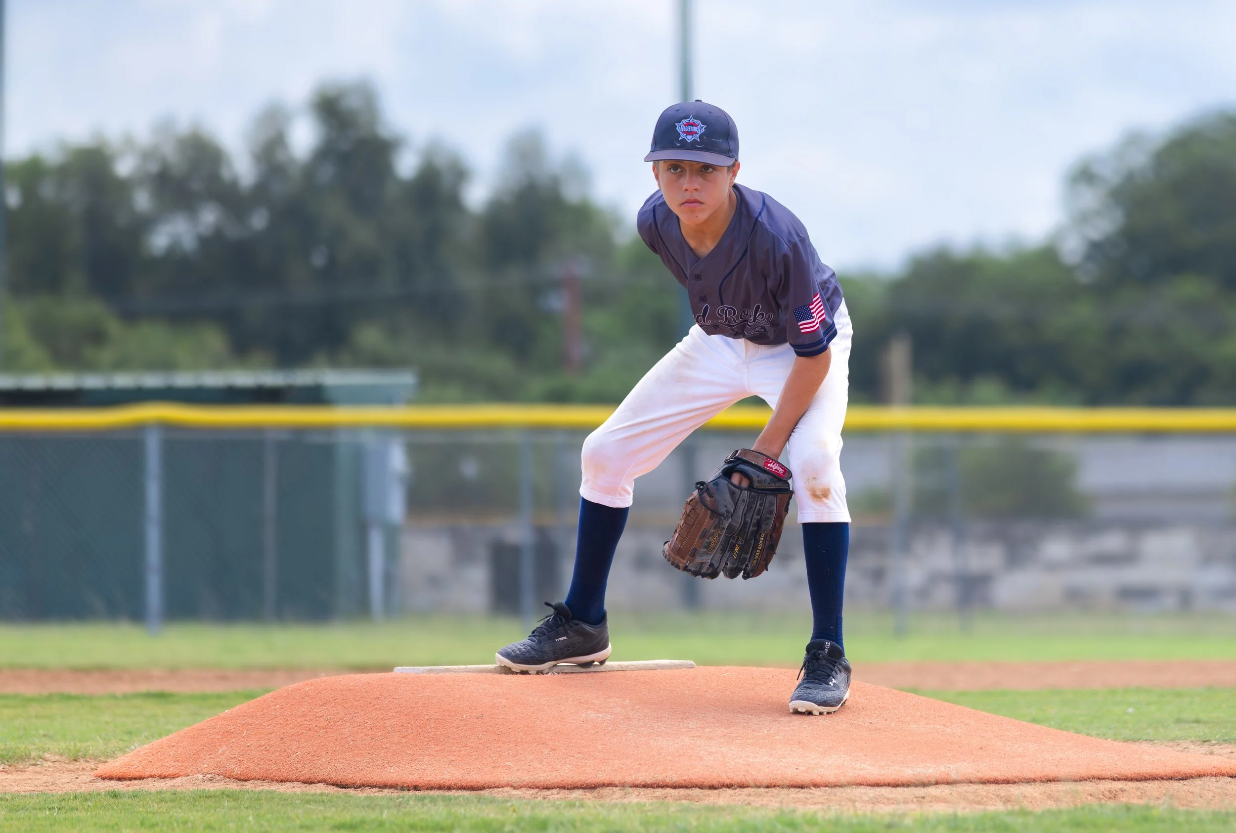 Young boy in a baseball uniform and cap, standing on the pitcher's mound on a baseball field, wearing a glove and preparing to pitch.